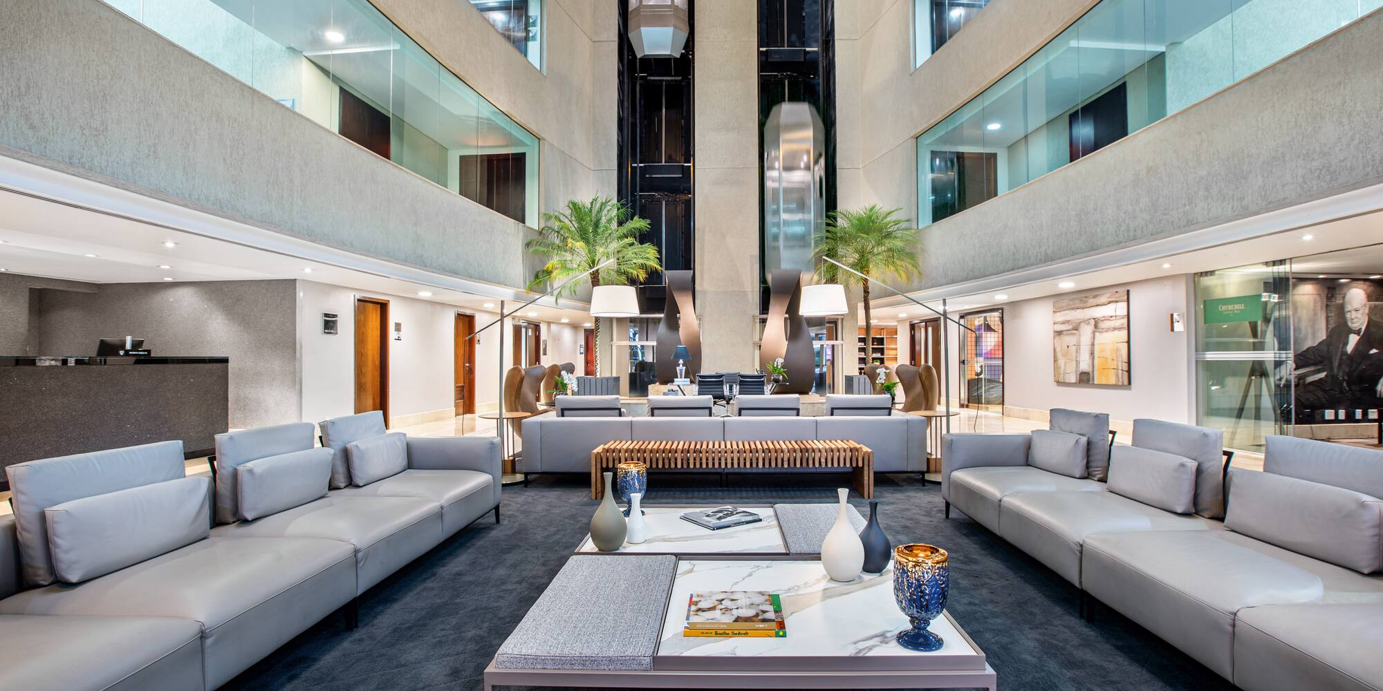 Modern hotel lobby with gray lounge sofas, marble tables, and glass elevators.