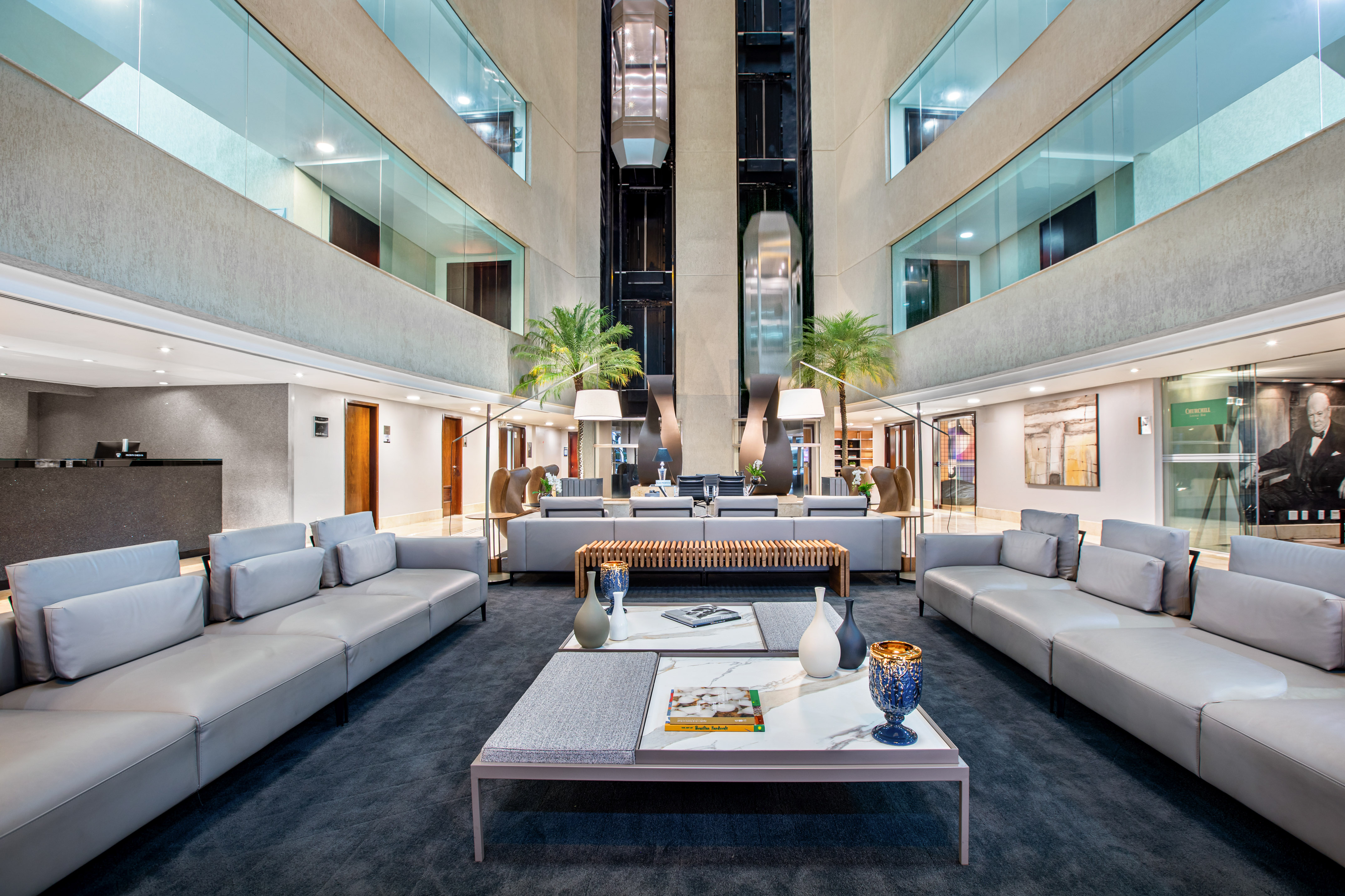 Modern hotel lobby with gray lounge sofas, marble tables, and glass elevators.