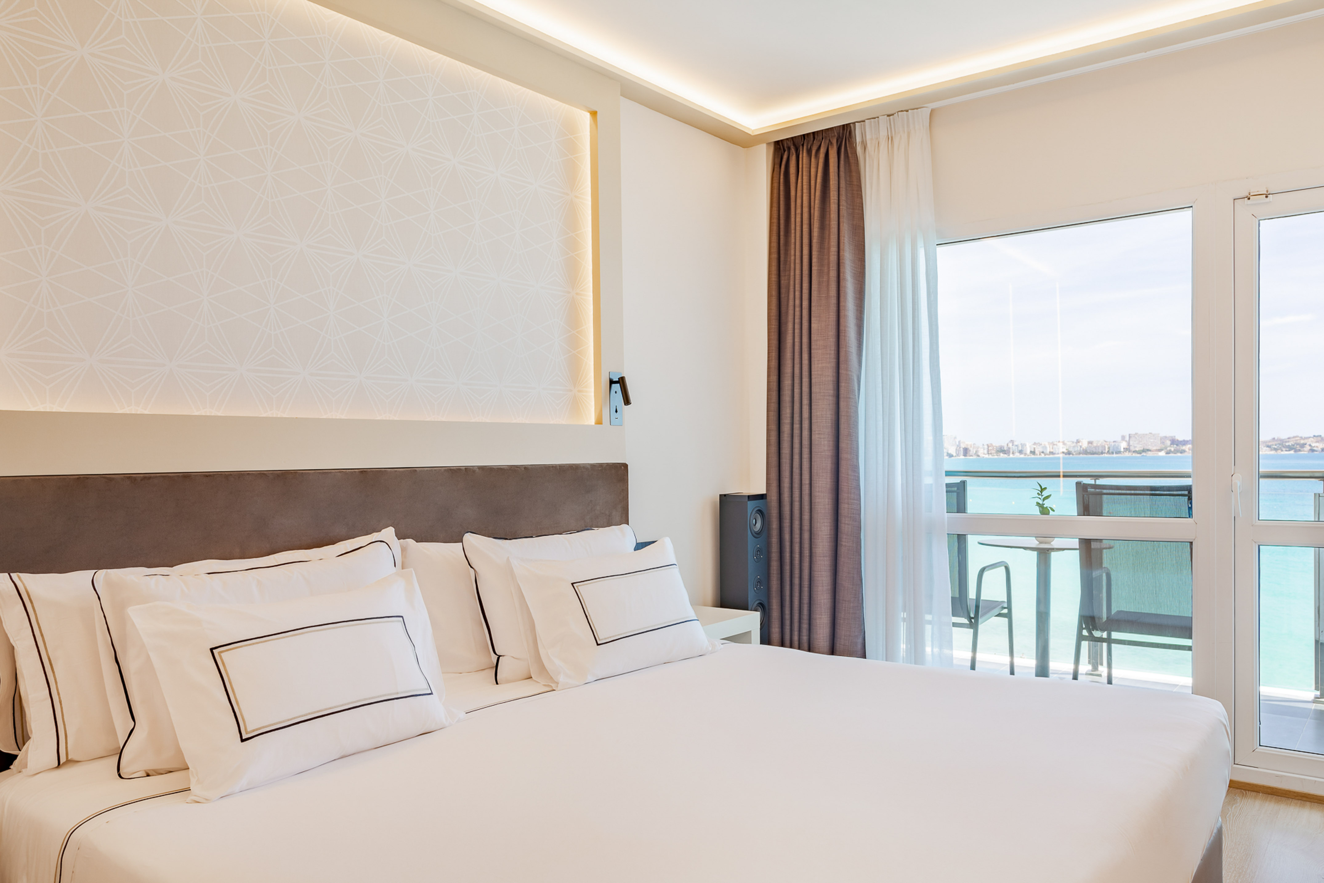 Modern hotel bedroom with white bed, patterned wall, and sea view balcony.