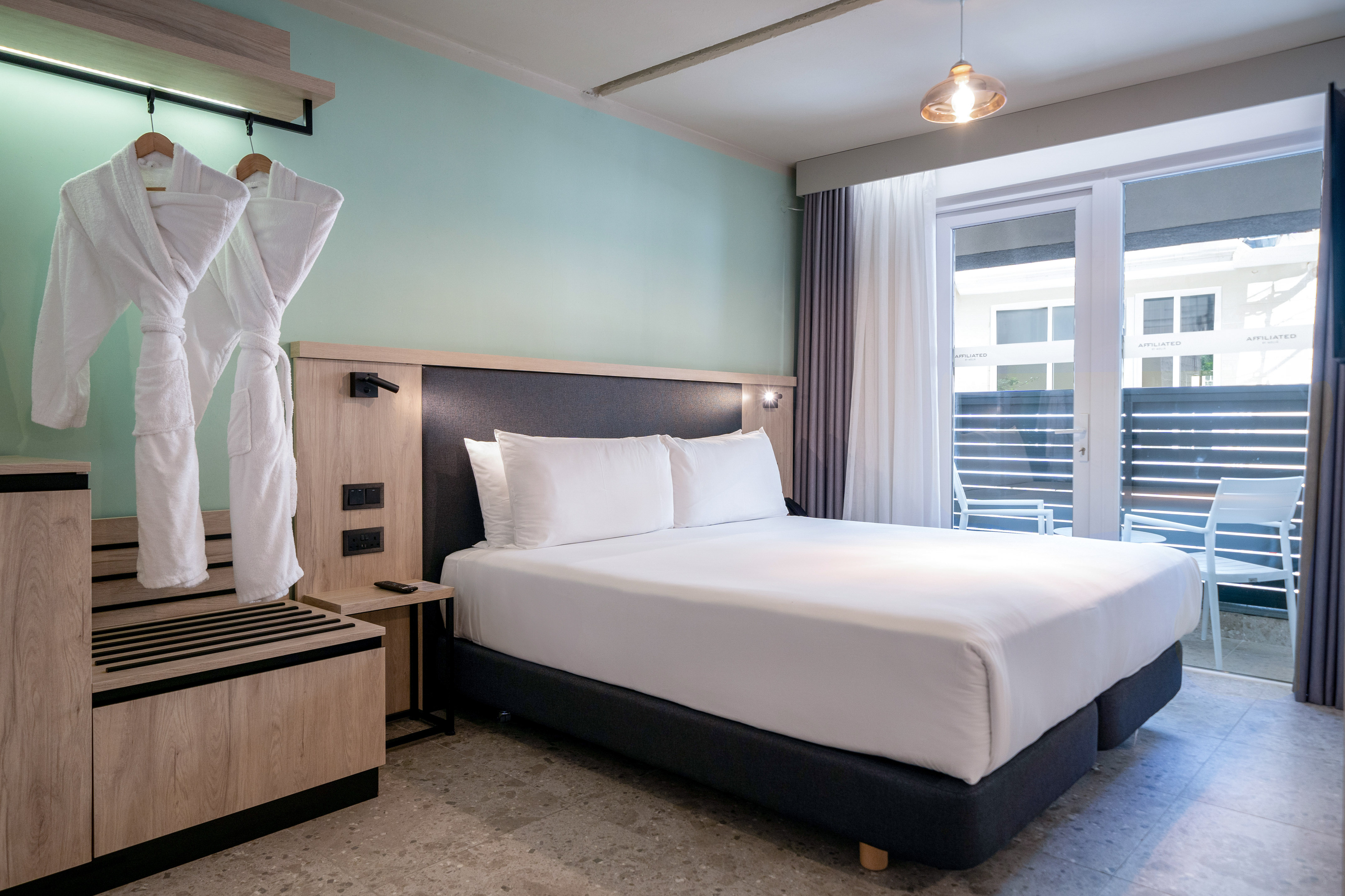 Modern hotel room with king bed, light blue wall, bathrobes, and balcony.