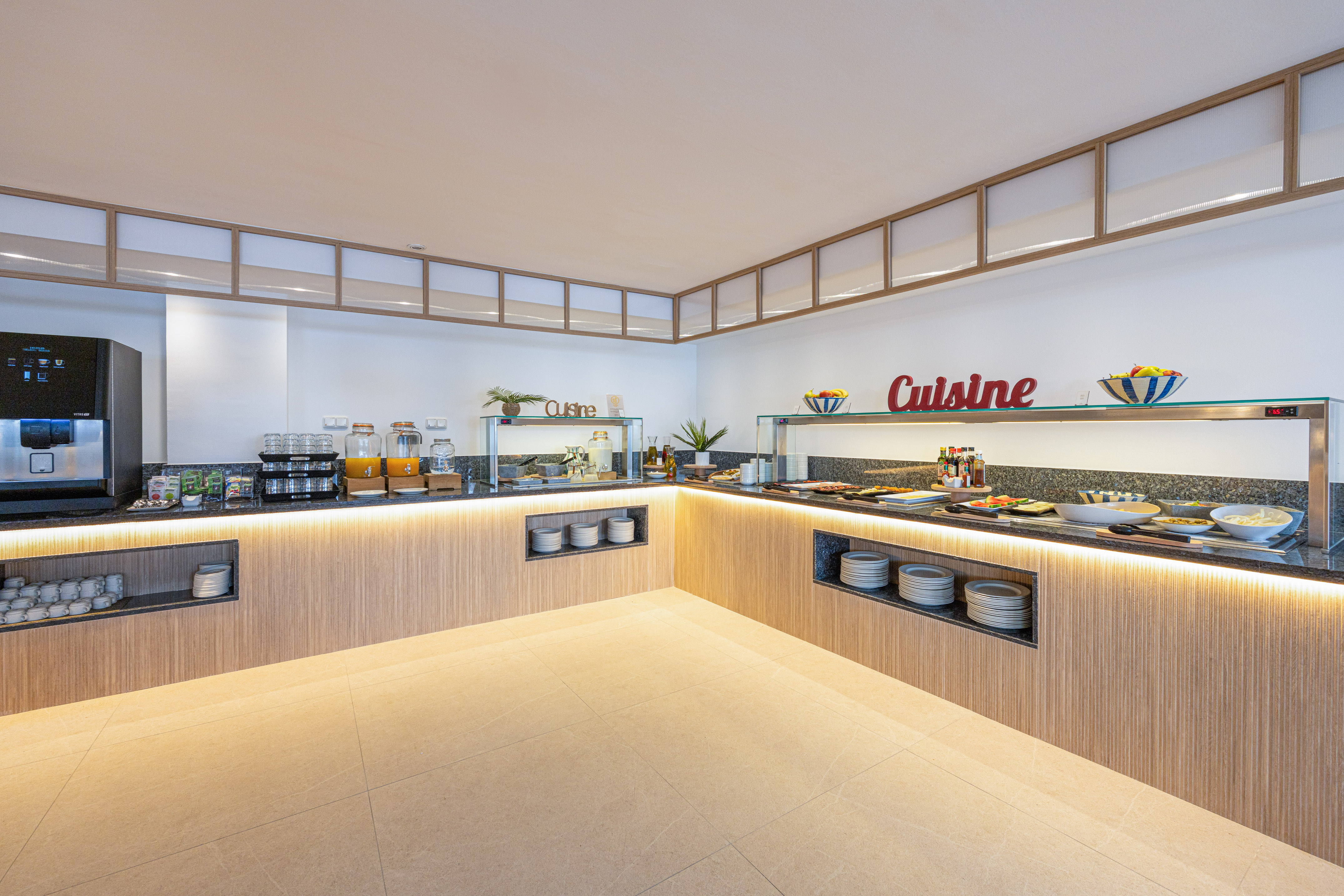 Bright hotel buffet with breakfast and "Cuisine" sign, juices, and coffee machine.