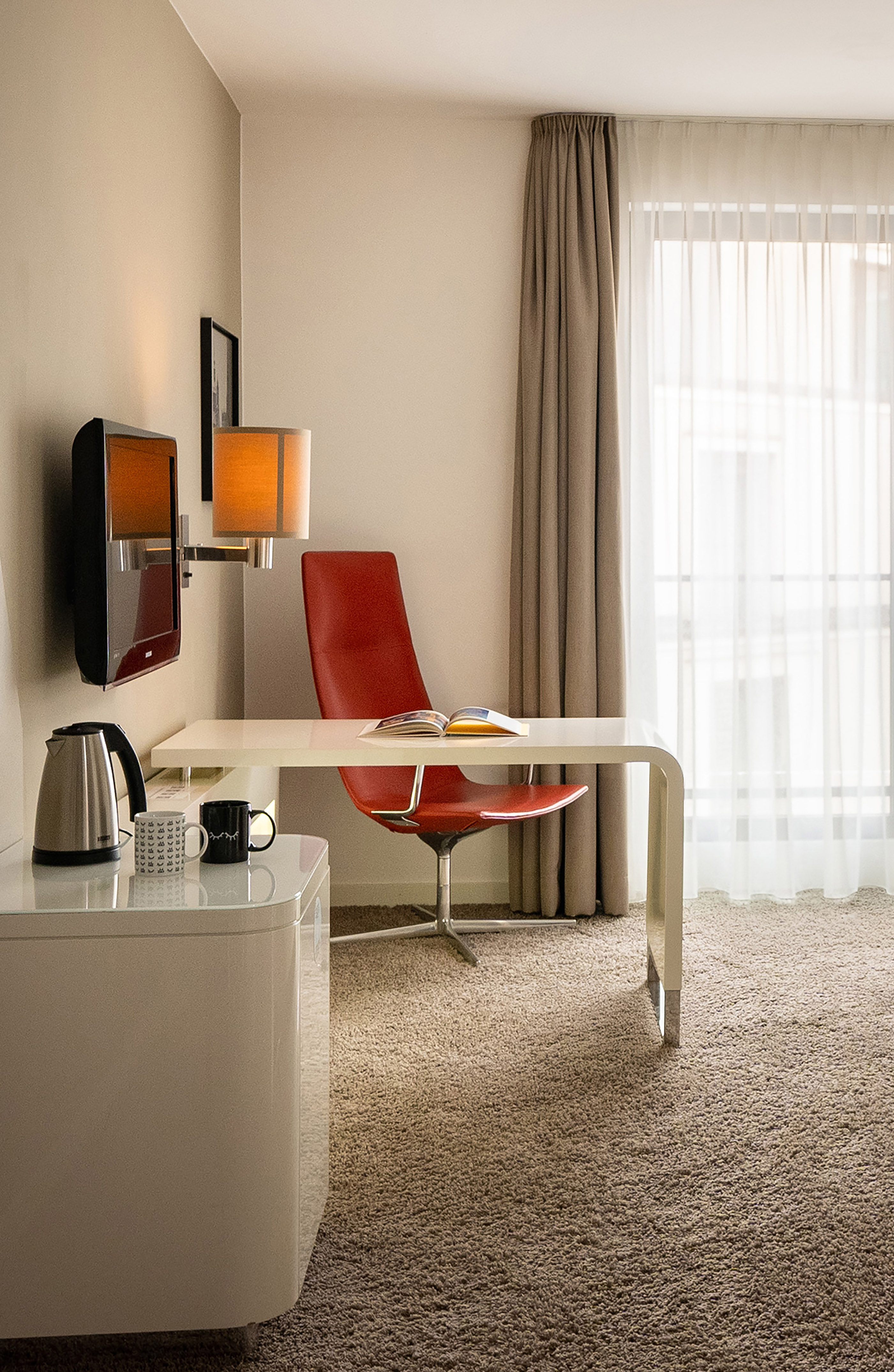 Contemporary hotel room workspace with red chair, white desk, and natural light.
