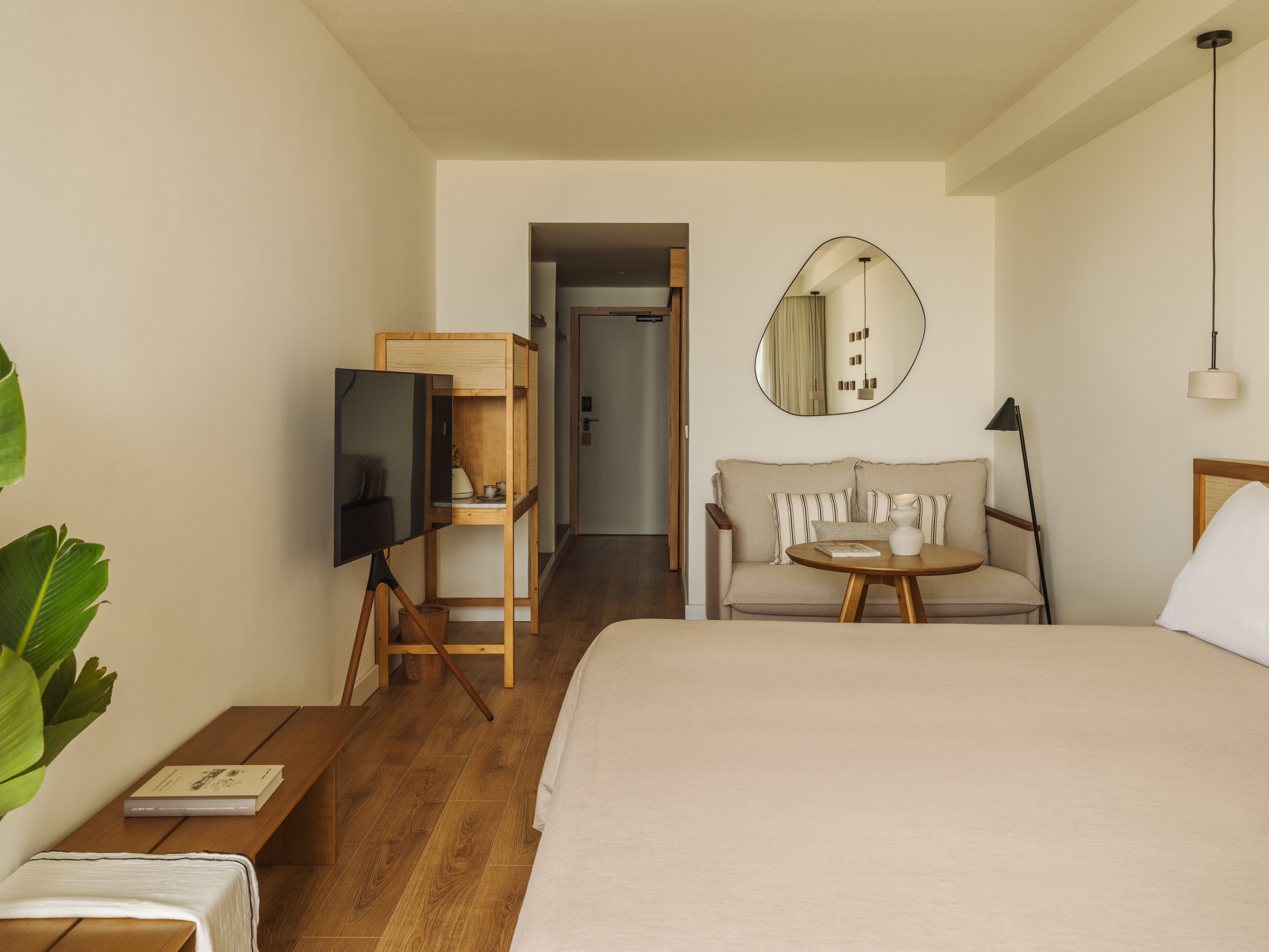 Modern hotel room with neutral decor, bed, sofa, TV, and wooden furniture.