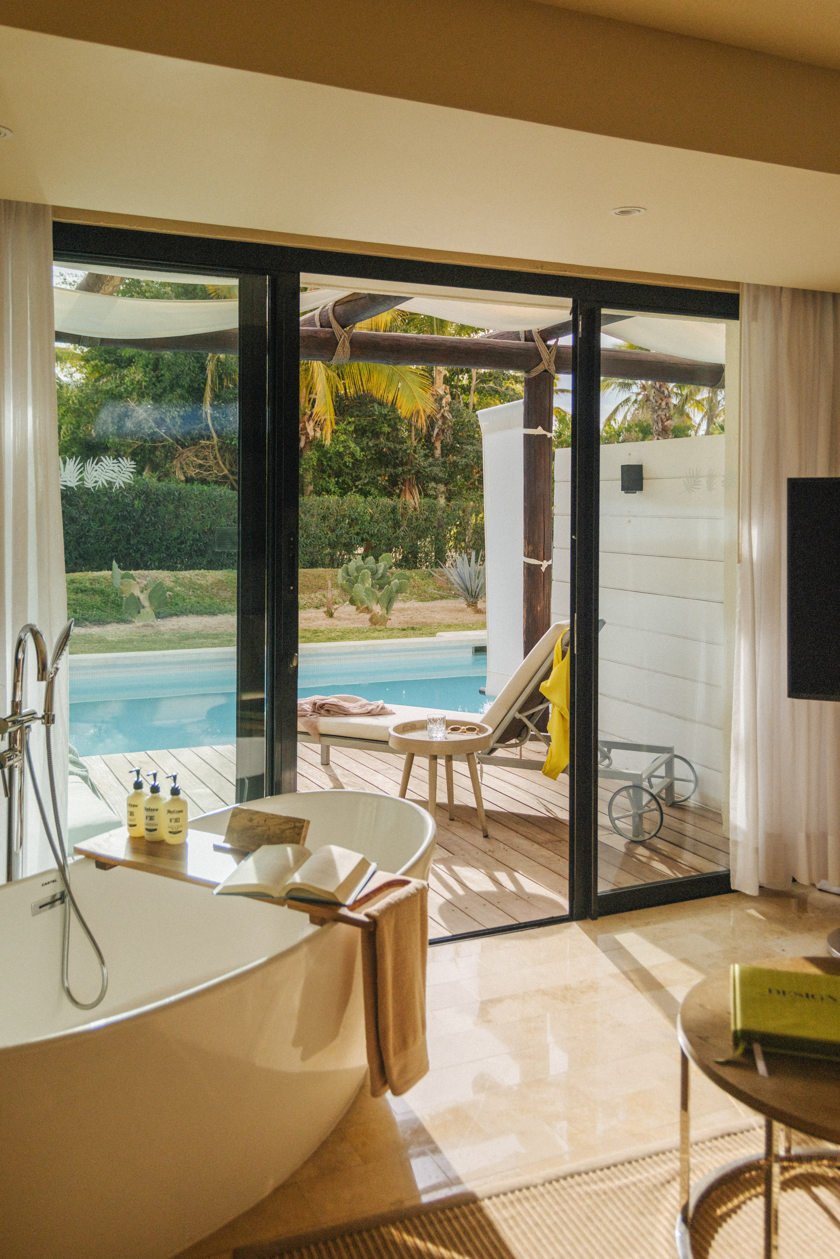 Luxury hotel bathroom with freestanding tub overlooking private pool & tropical deck.