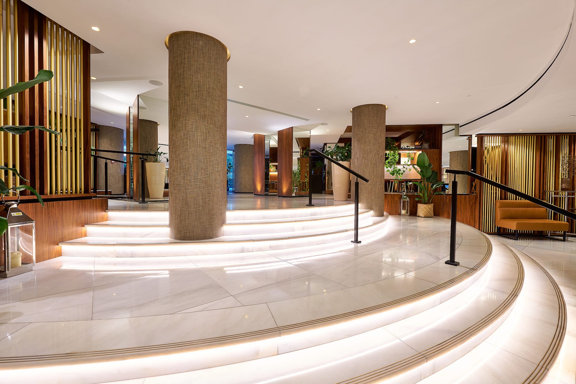 Luxury hotel lobby with illuminated marble stairs, textured columns, and plants.