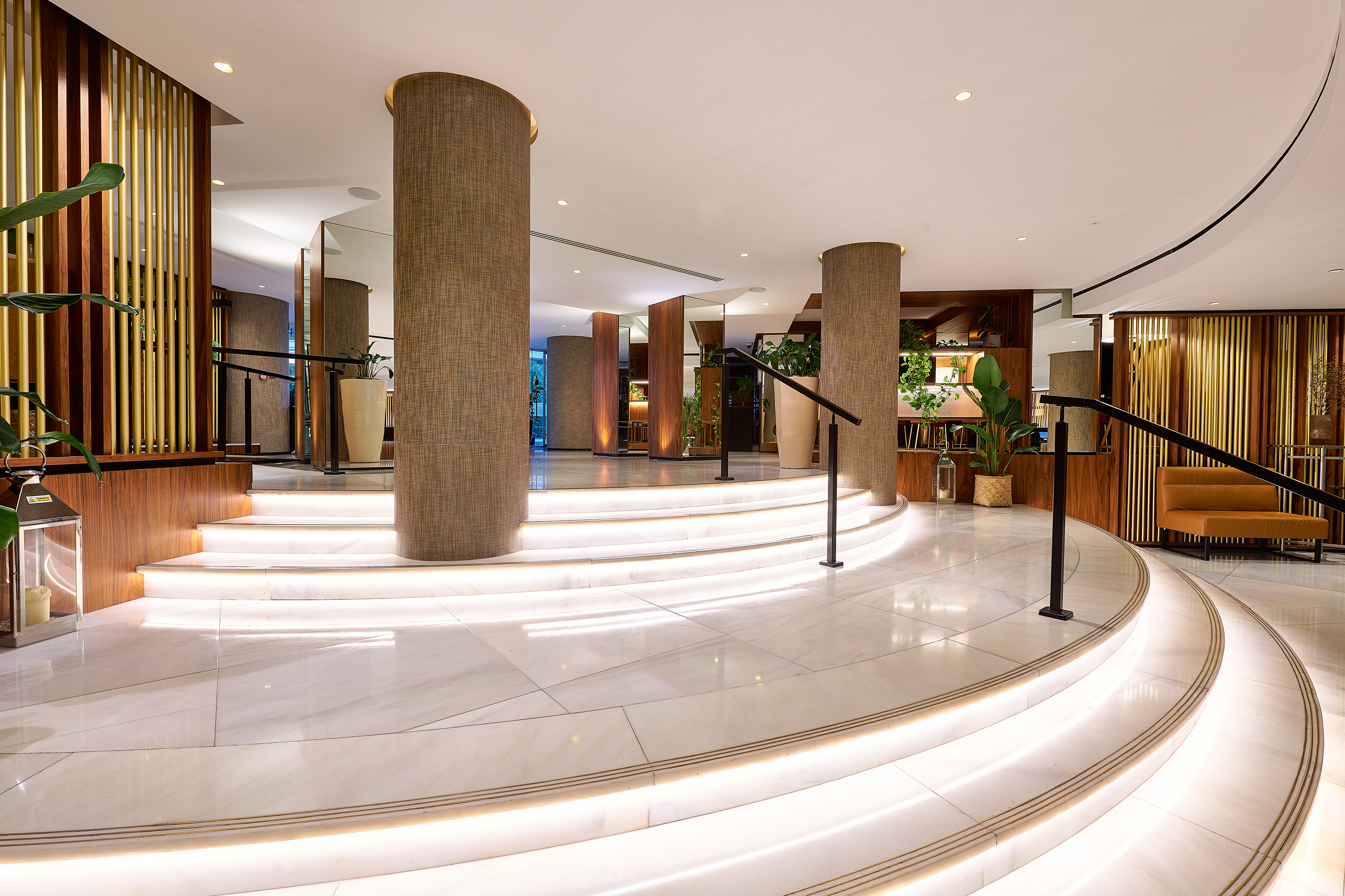 Luxury hotel lobby with illuminated marble stairs, textured columns, and plants.