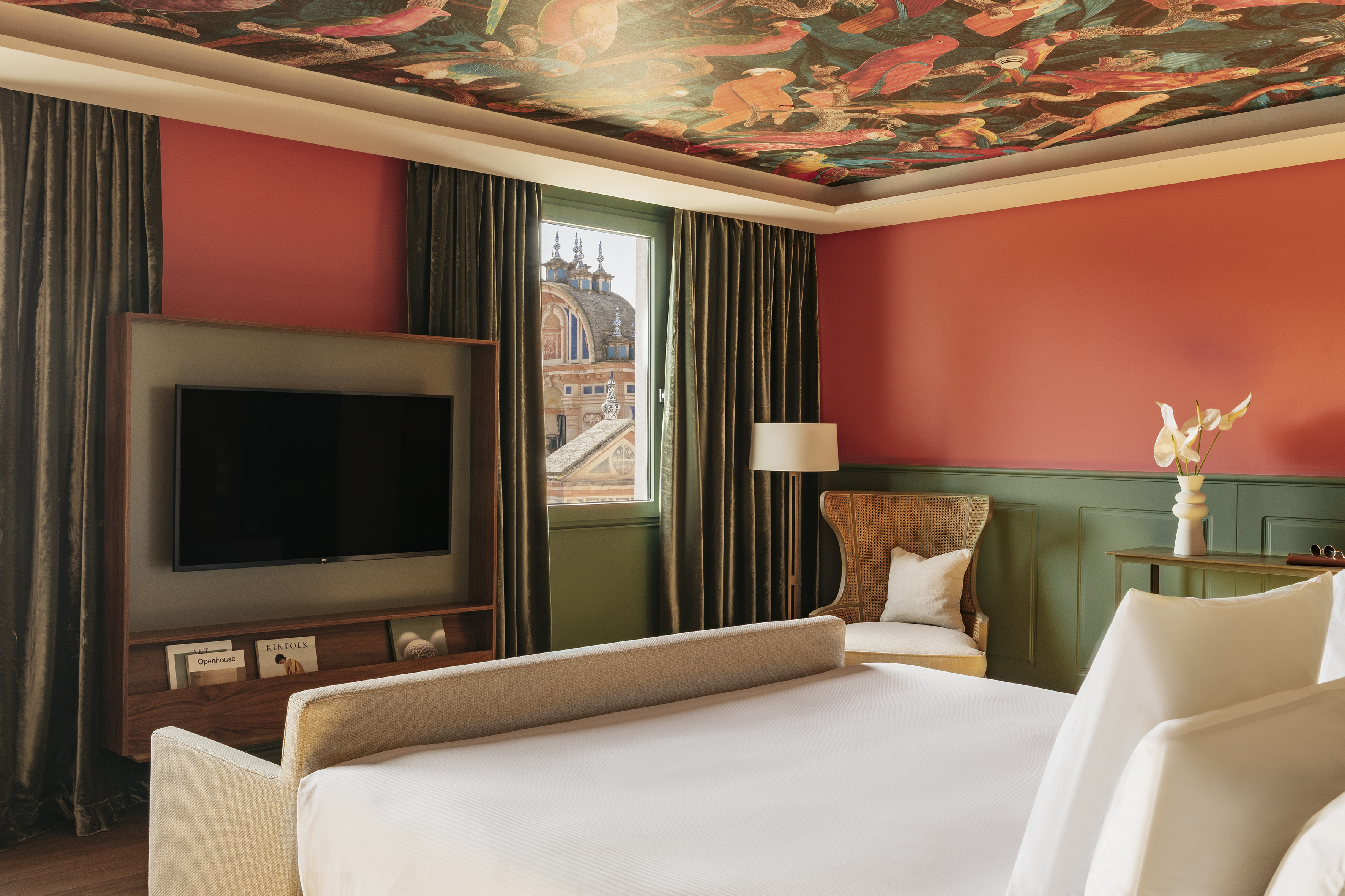Vibrant hotel bedroom with coral walls, bird mural ceiling, and historic view.