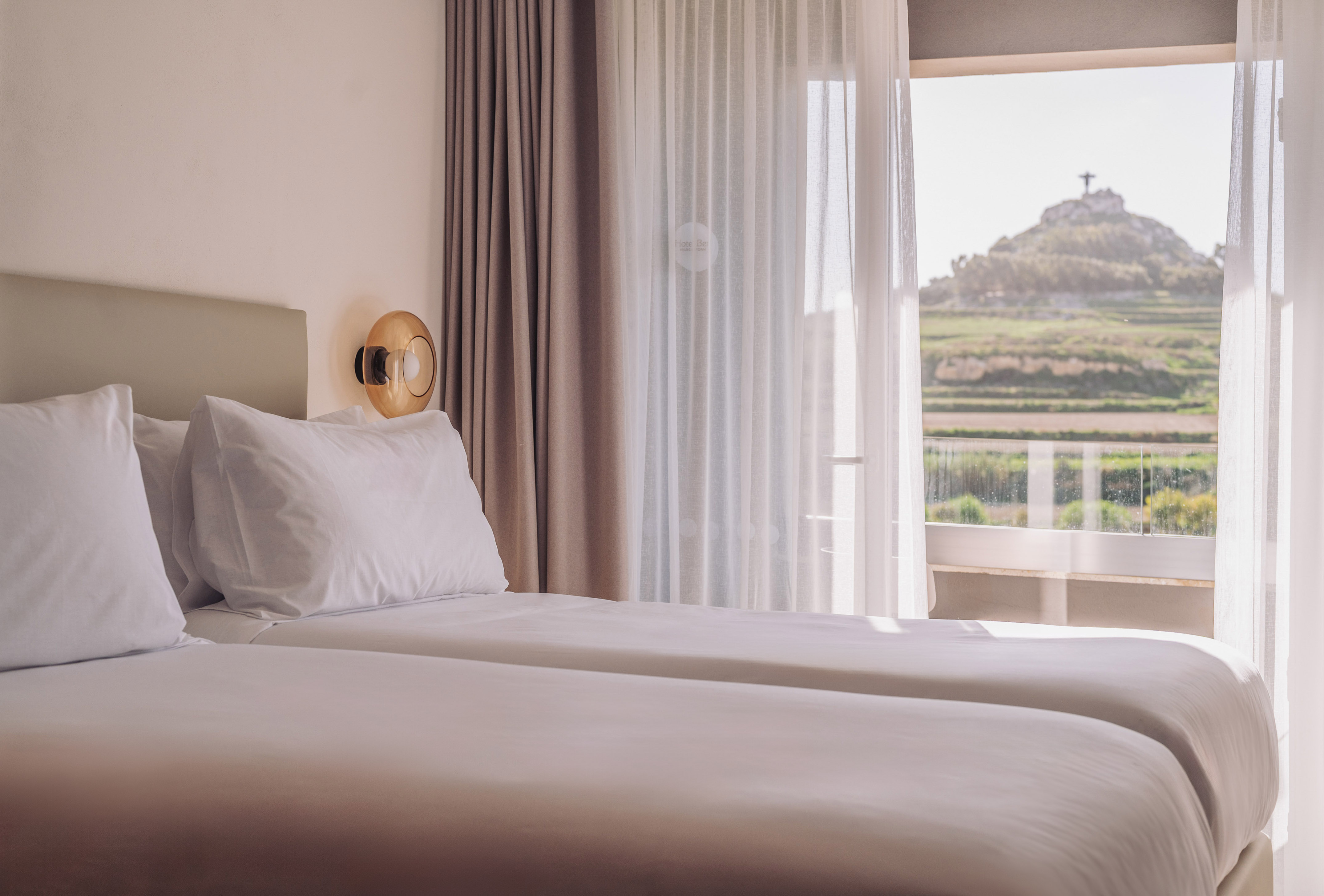 Bright hotel room with twin beds, mountain view, and hilltop statue through window.