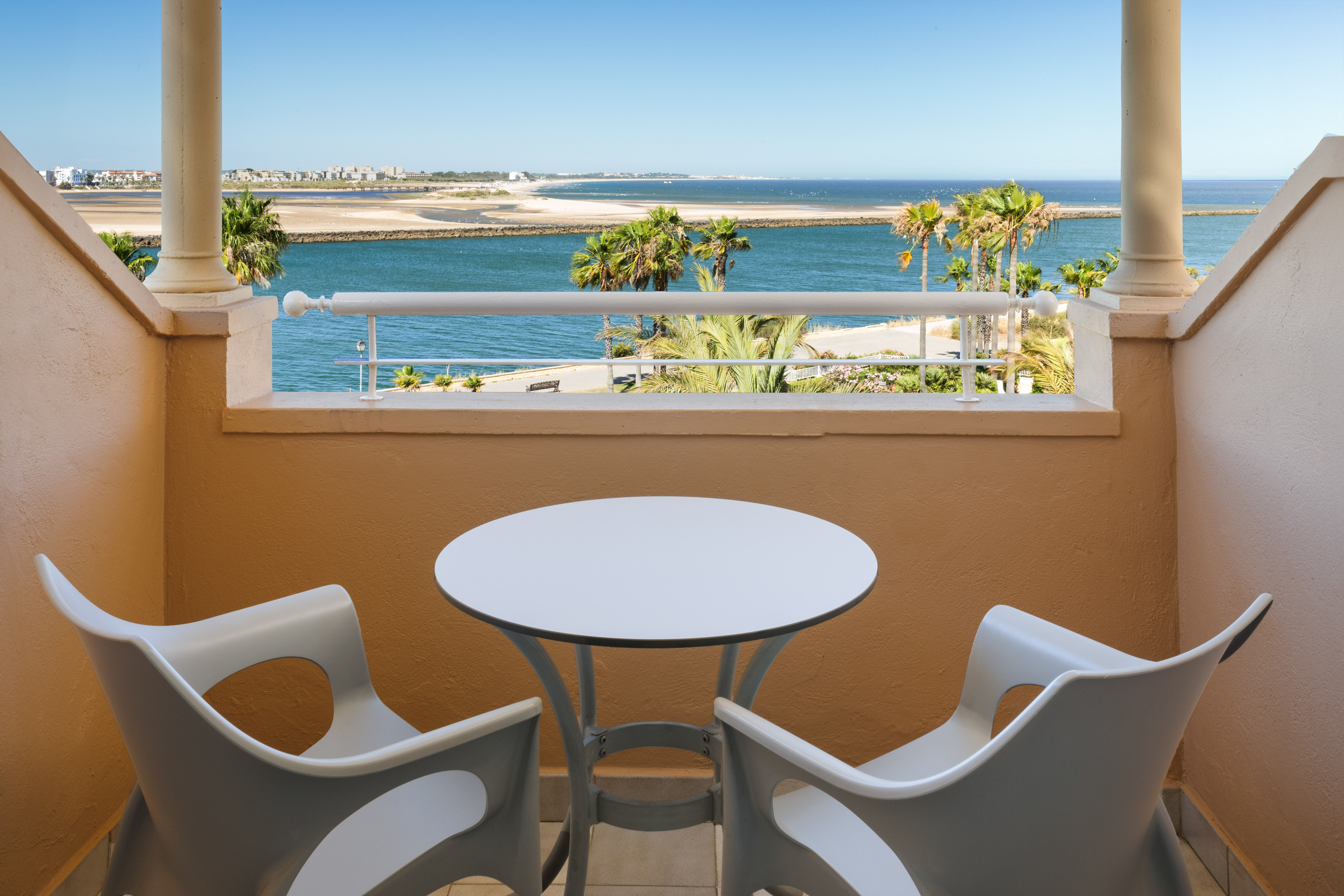 Coastal balcony with ocean, palm trees, beach. White outdoor furniture.