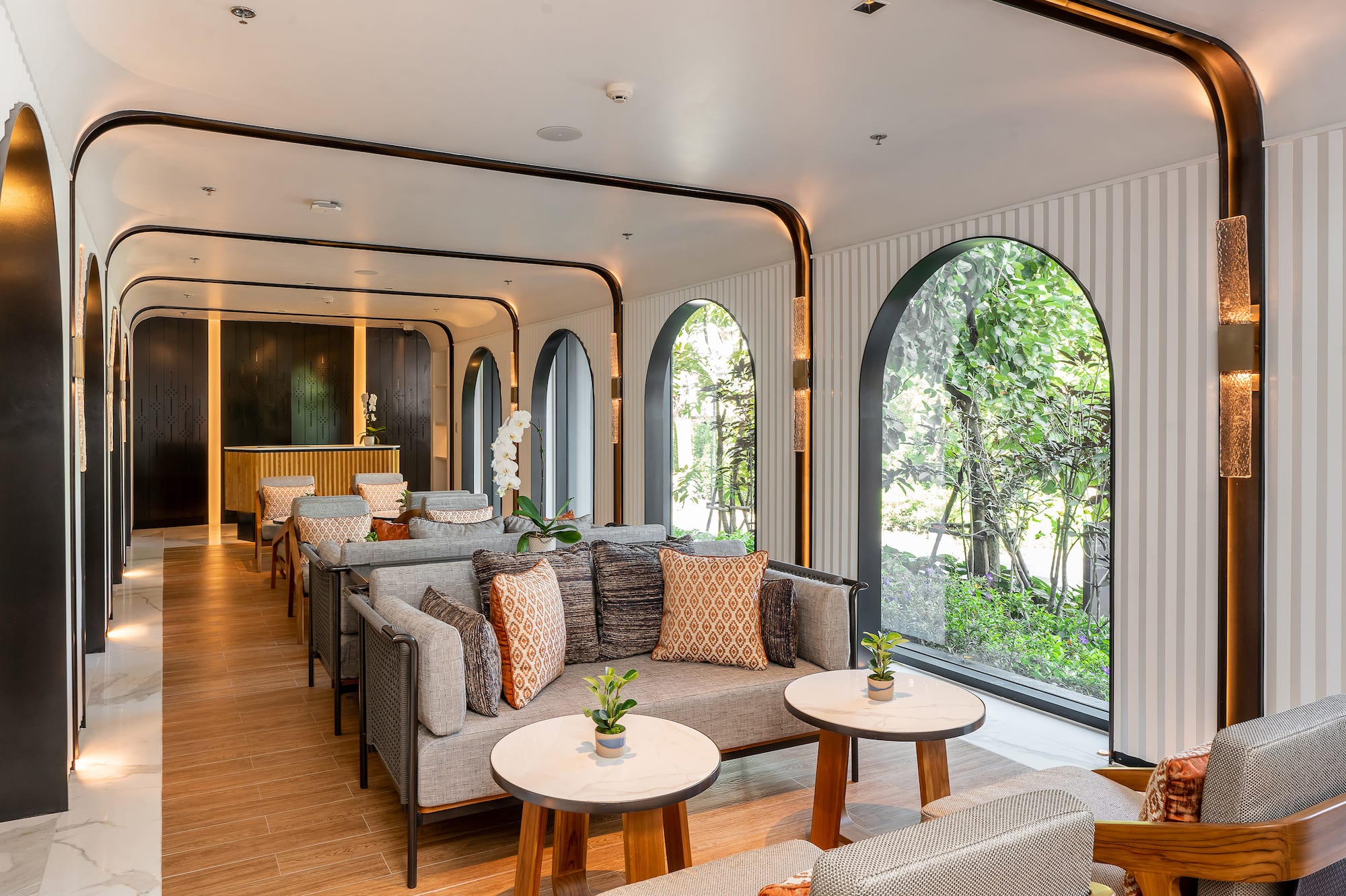 Modern hotel lounge with grey sofas, arched windows, and lush garden views.