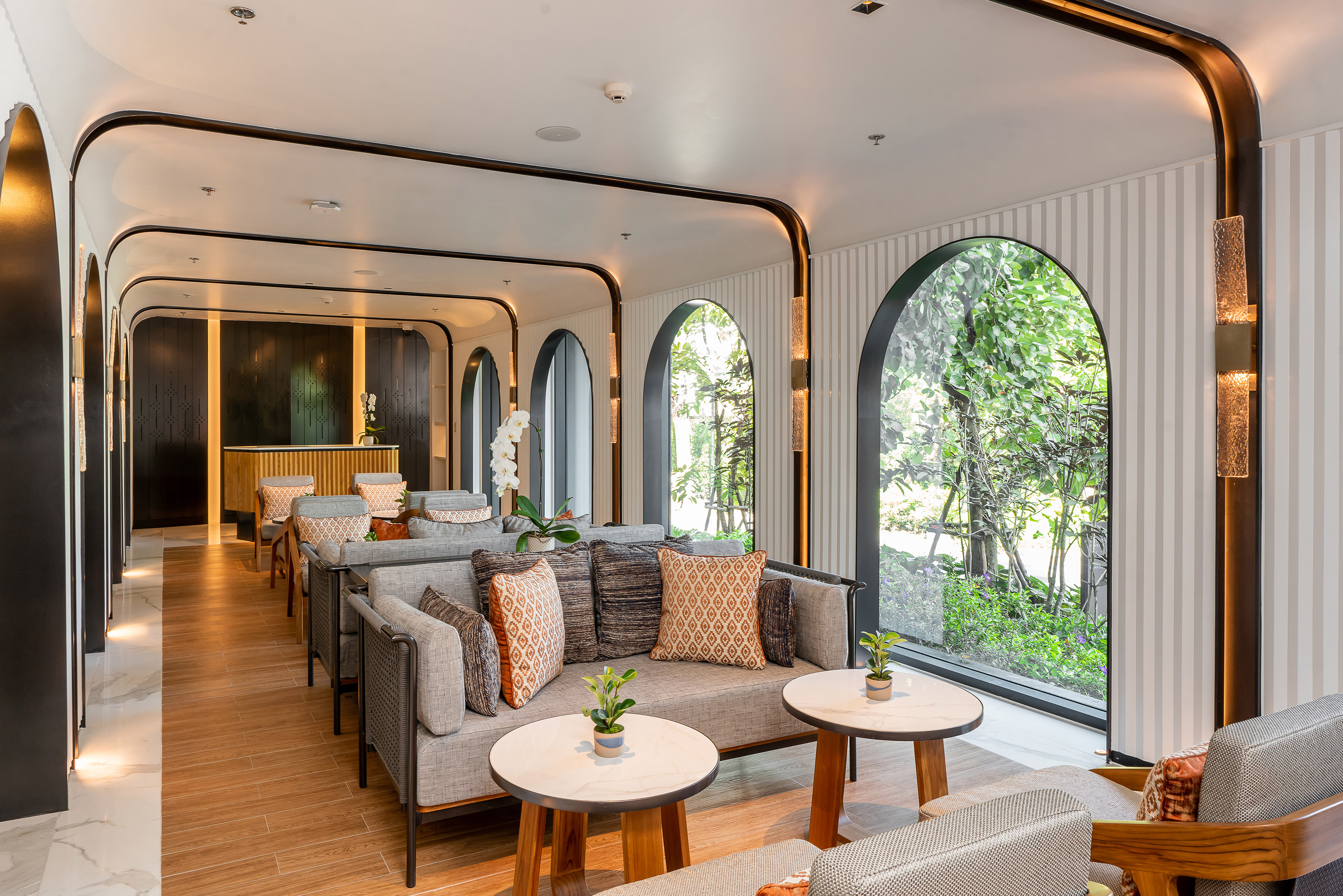 Modern hotel lounge with grey sofas, arched windows, and lush garden views.