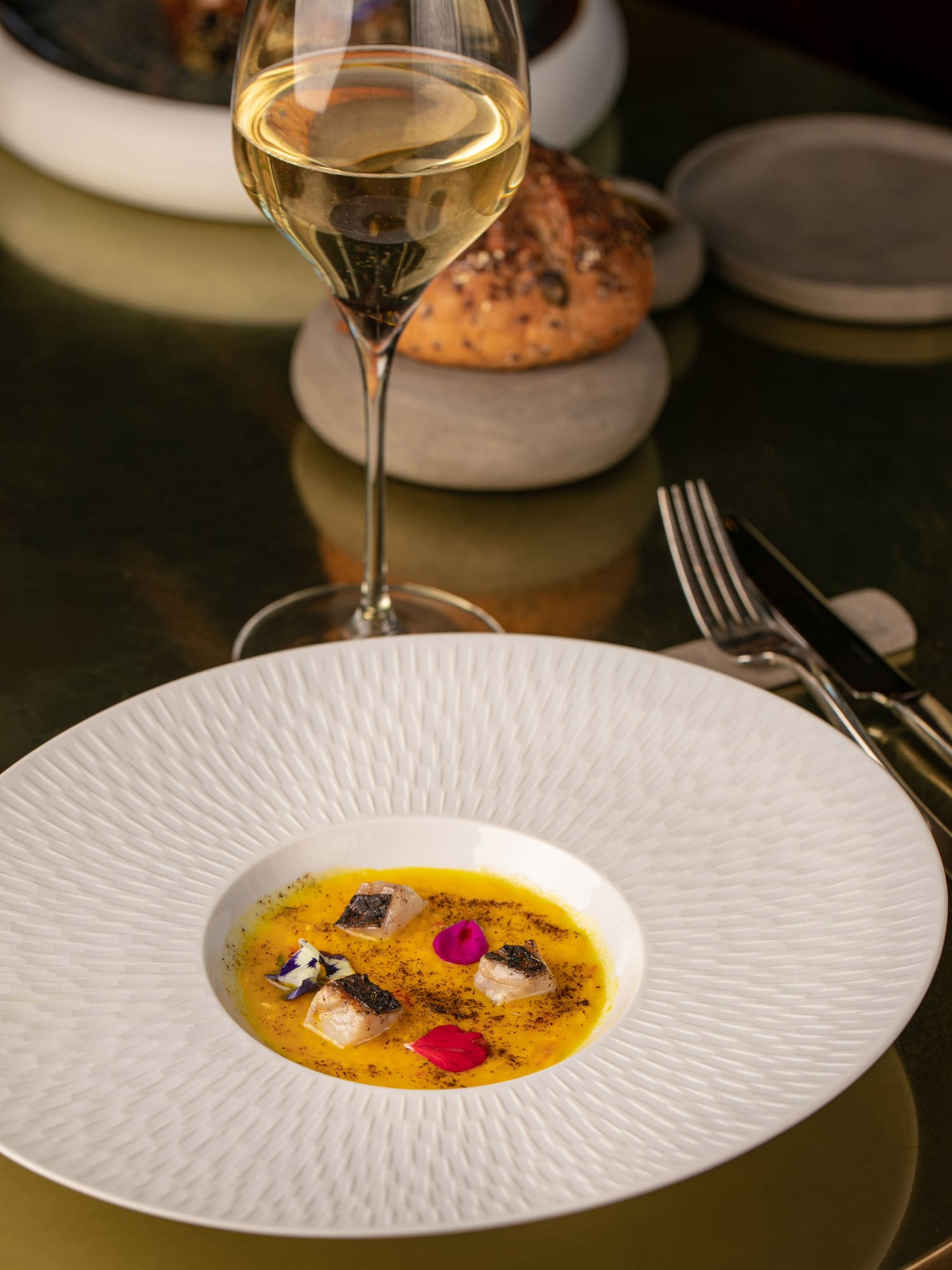 Elegant fish dish in yellow puree, garnished with flowers, white wine, and bread.