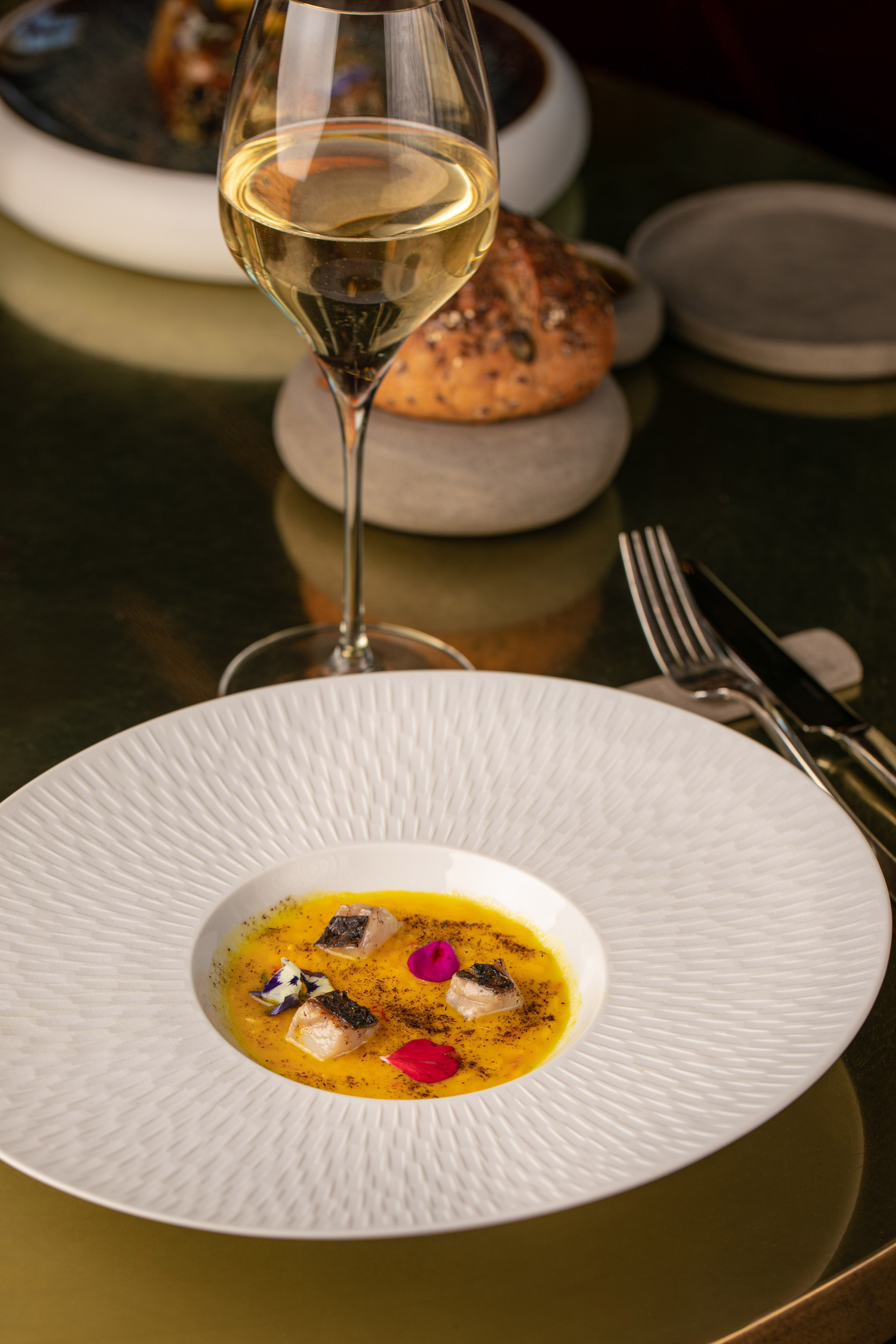 Elegant fish dish in yellow puree, garnished with flowers, white wine, and bread.