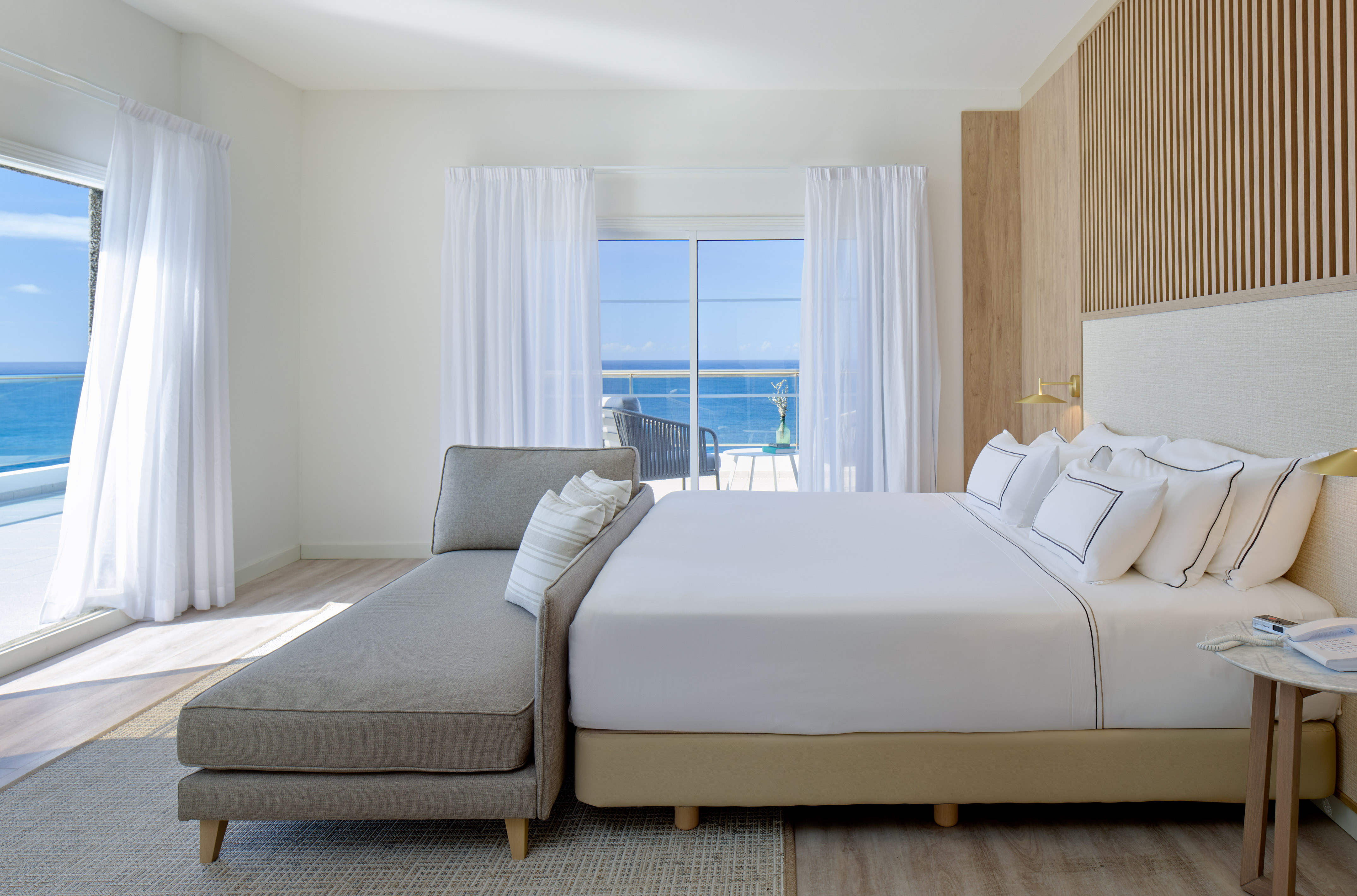 Modern hotel bedroom with ocean view, white bed, grey chaise, balcony.