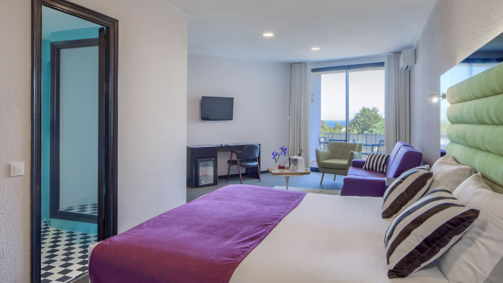 Modern hotel suite with purple bed, living area, workspace, and ocean view balcony.