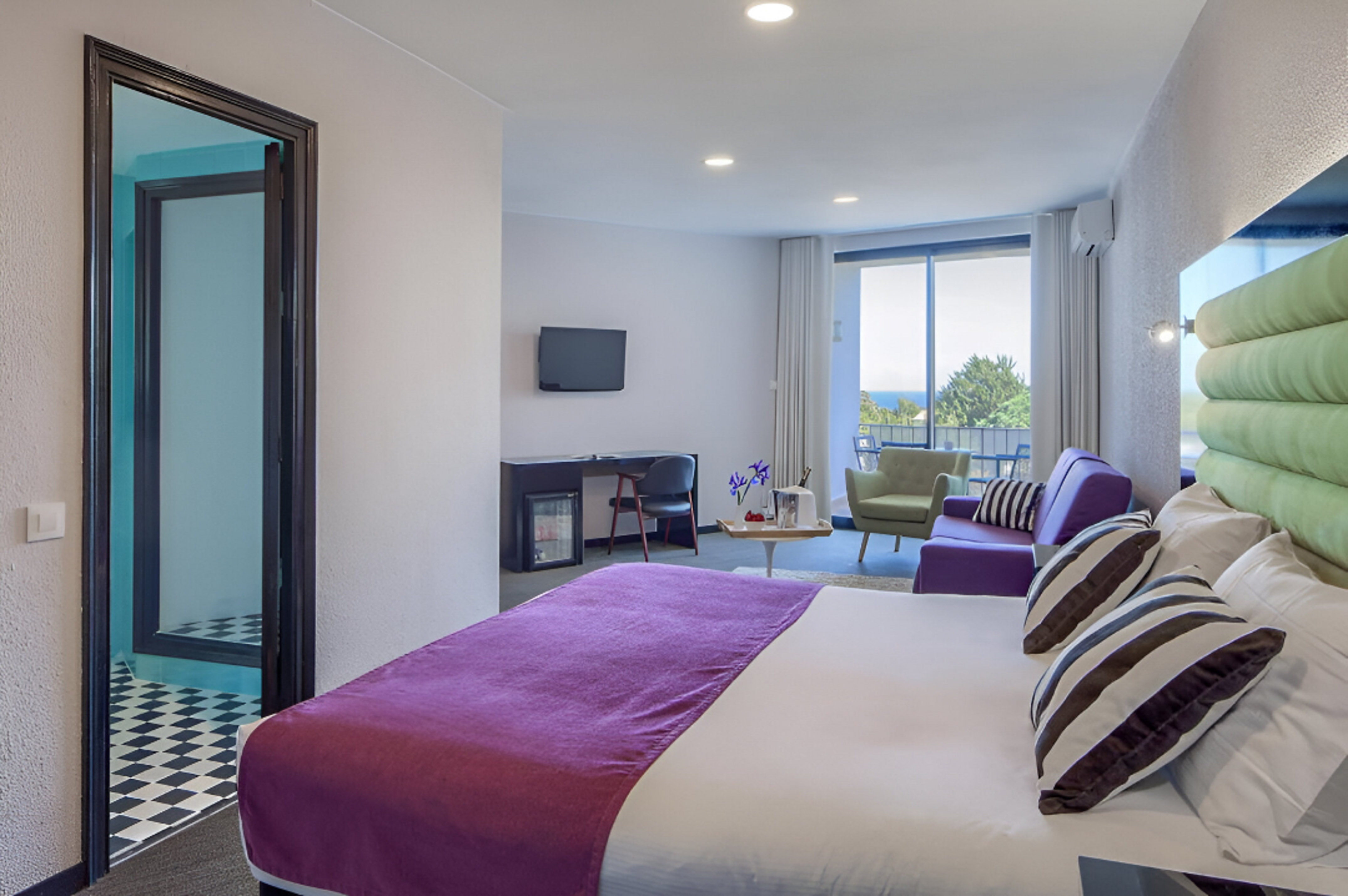 Modern hotel suite with purple bed, living area, workspace, and ocean view balcony.