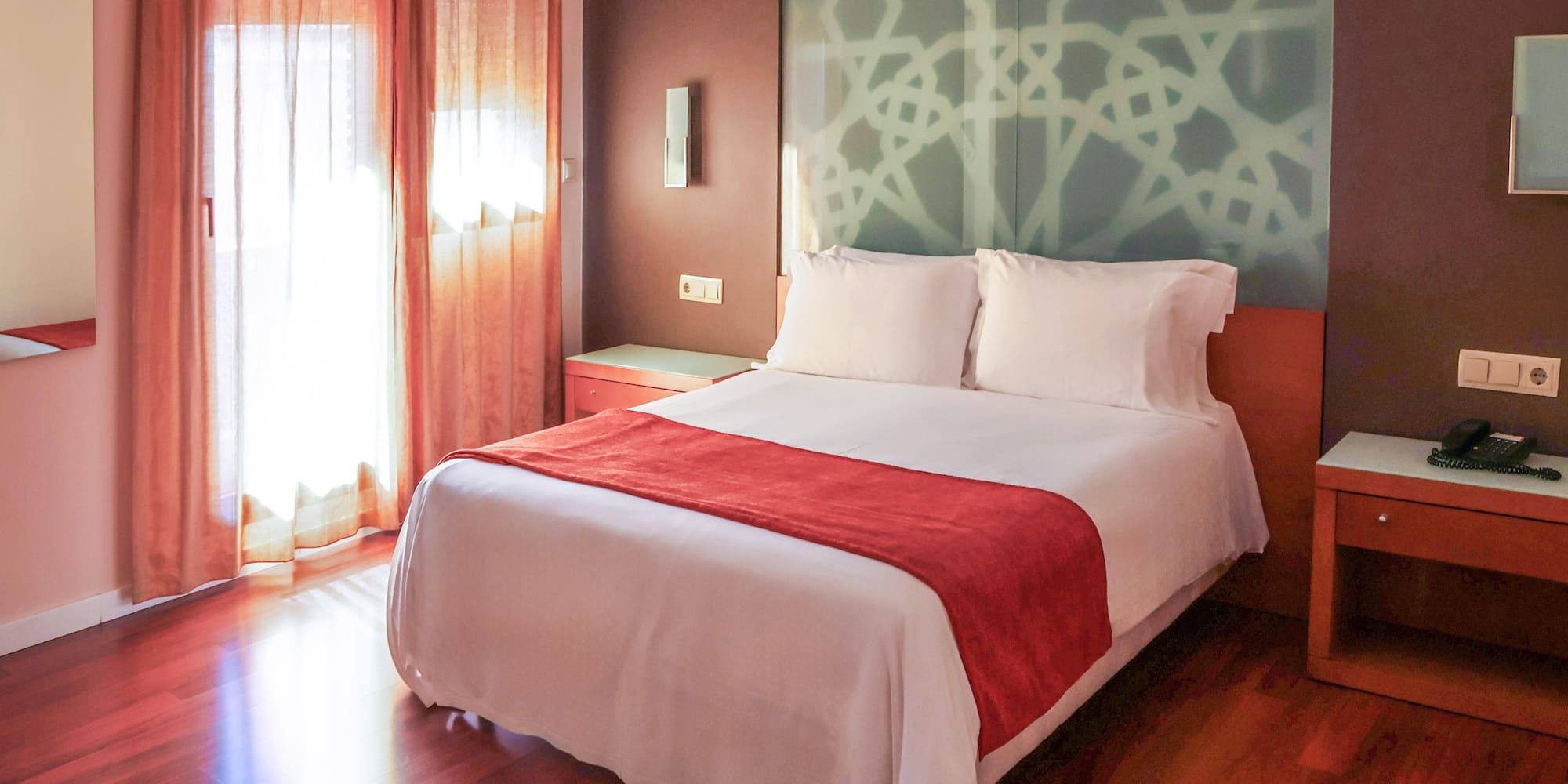 Hotel bedroom: white bed, red runner, patterned headboard, sunlit window.