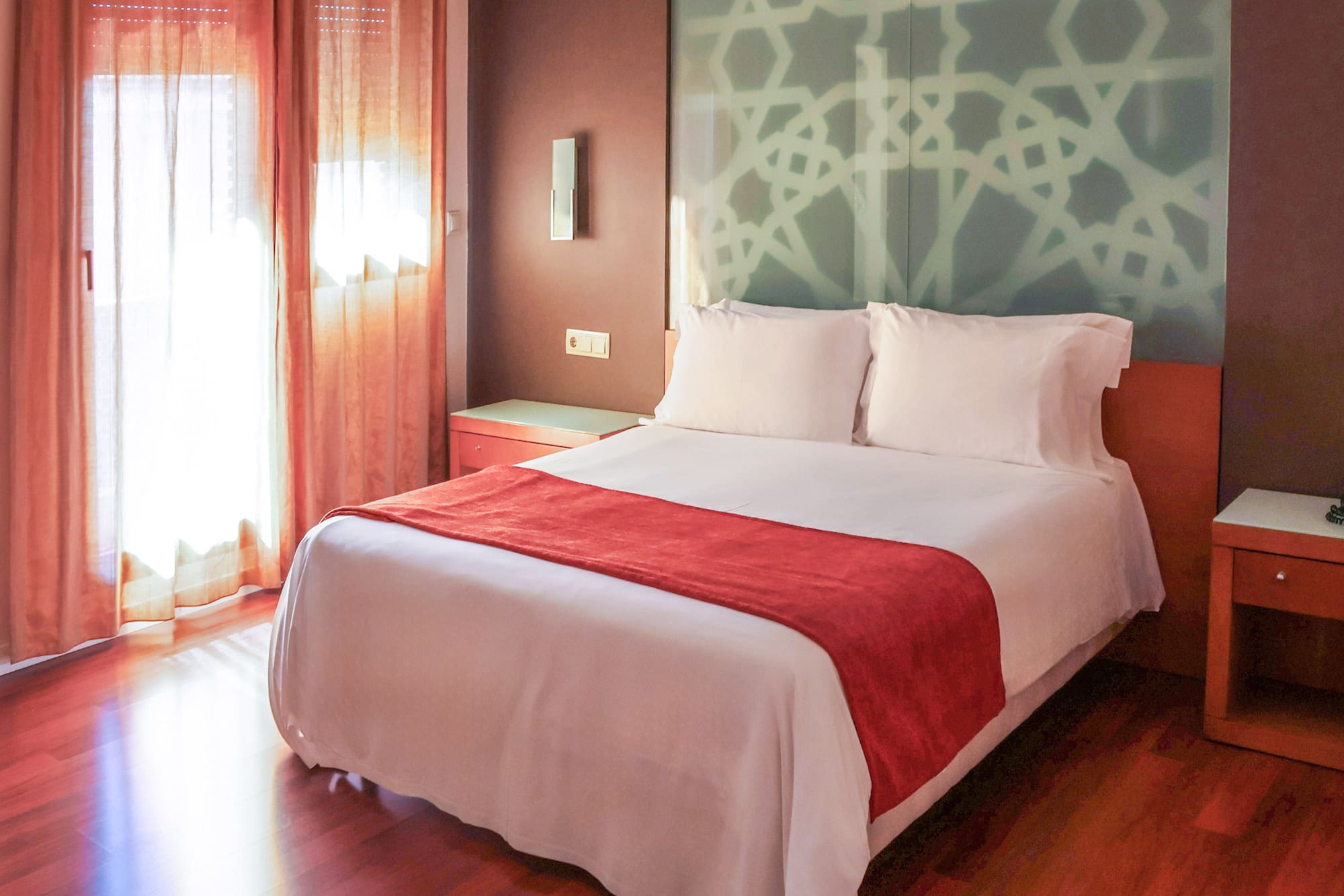 Hotel bedroom: white bed, red runner, patterned headboard, sunlit window.