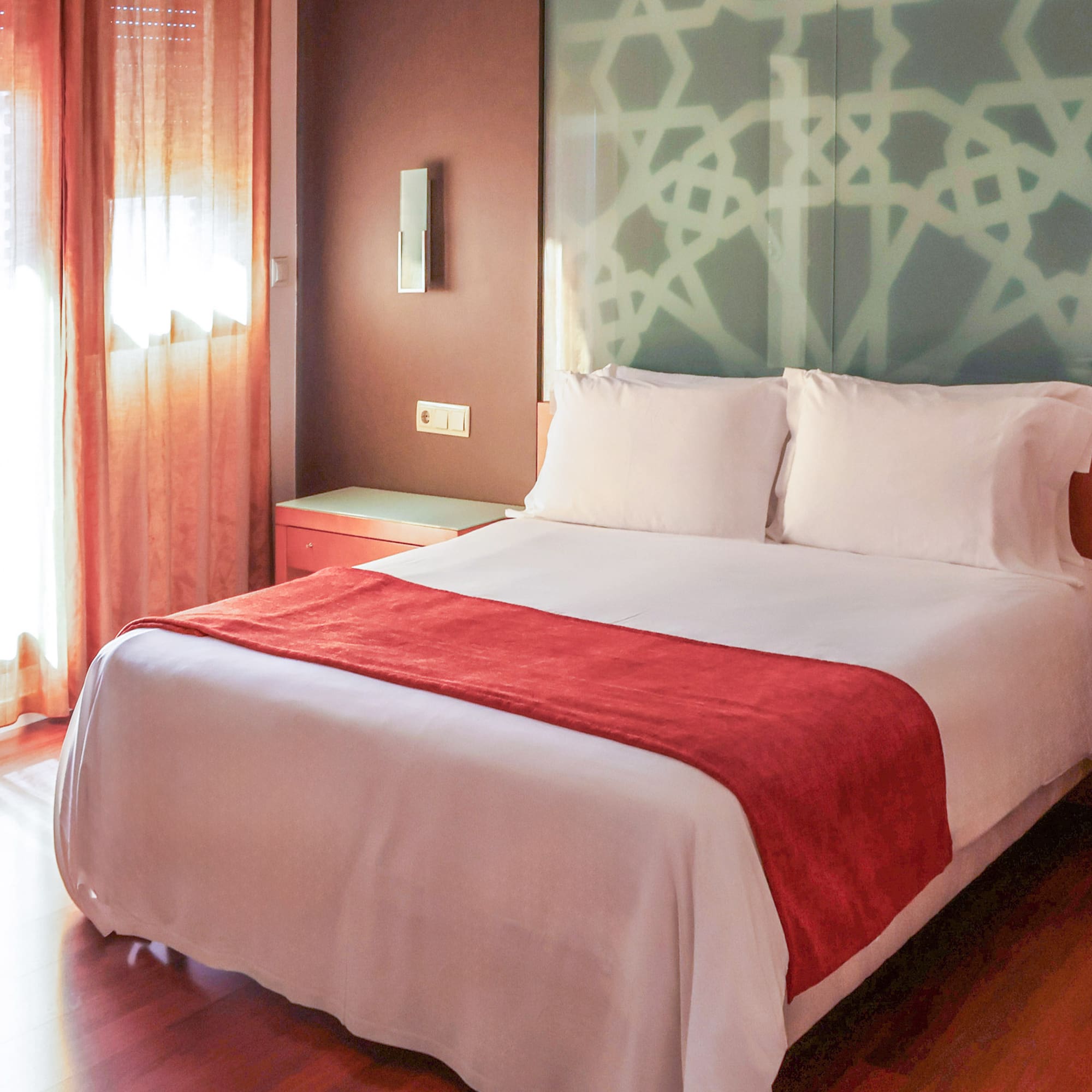 Hotel bedroom: white bed, red runner, patterned headboard, sunlit window.