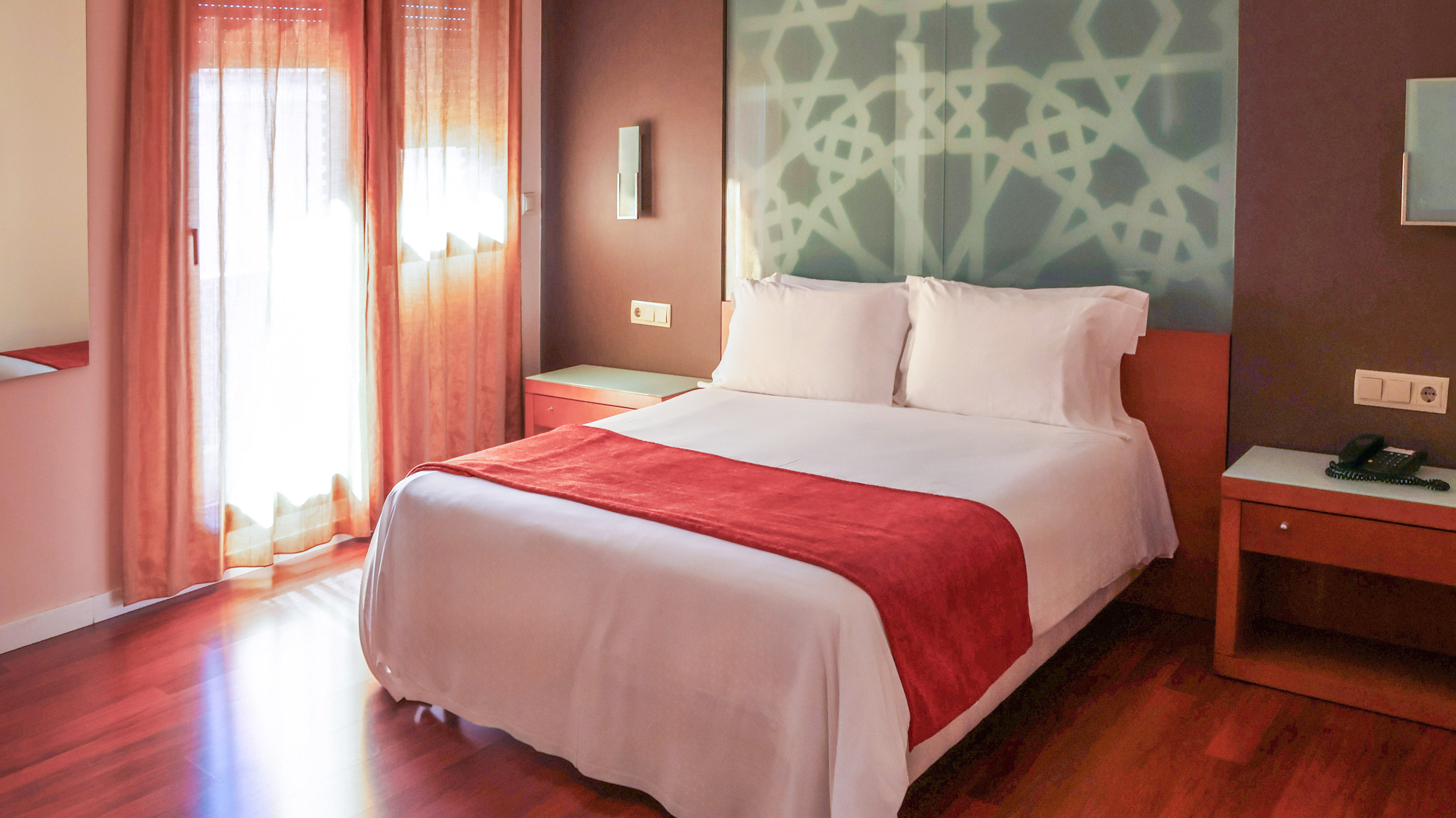 Hotel bedroom: white bed, red runner, patterned headboard, sunlit window.