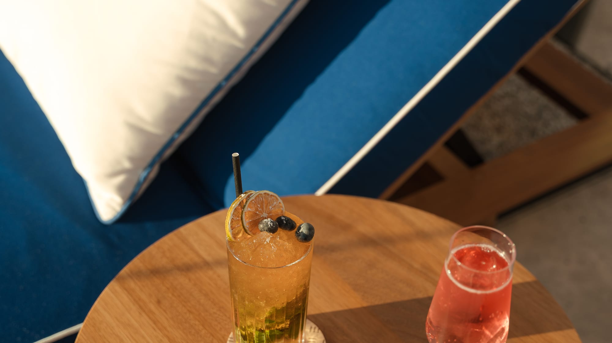 Iced cocktail and rosé on wooden table by a blue lounge chair outdoors.