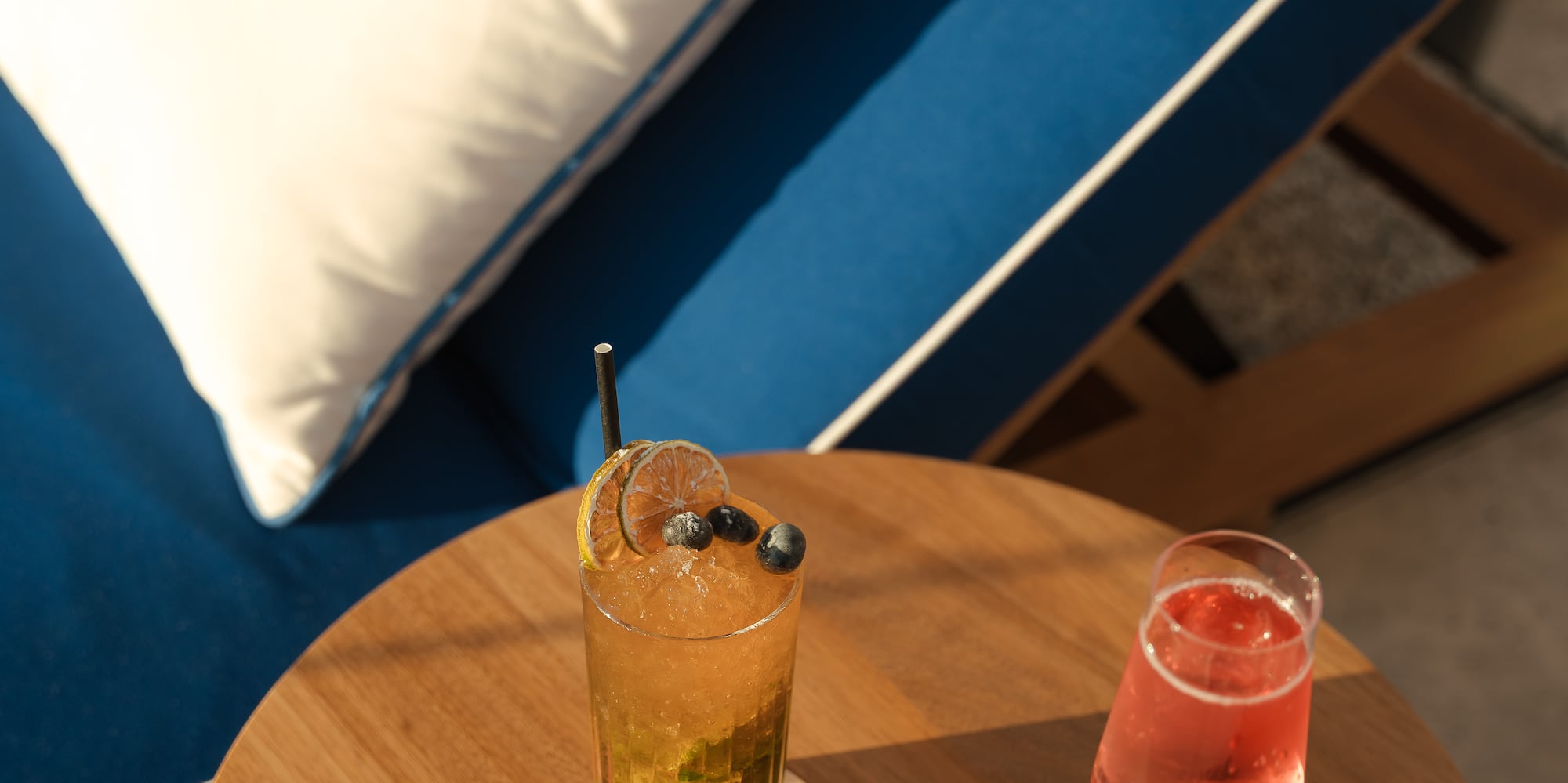 Iced cocktail and rosé on wooden table by a blue lounge chair outdoors.