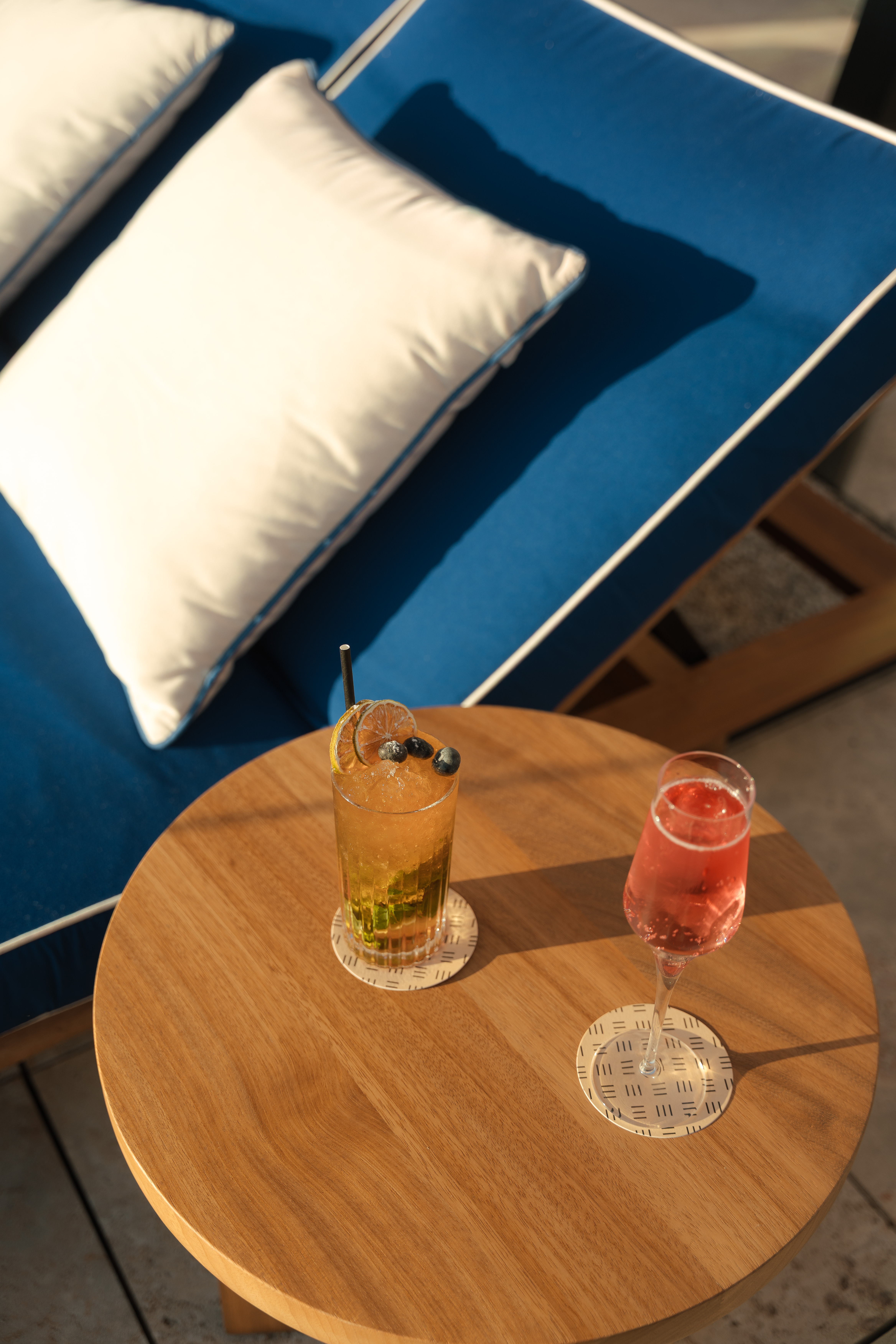 Iced cocktail and rosé on wooden table by a blue lounge chair outdoors.