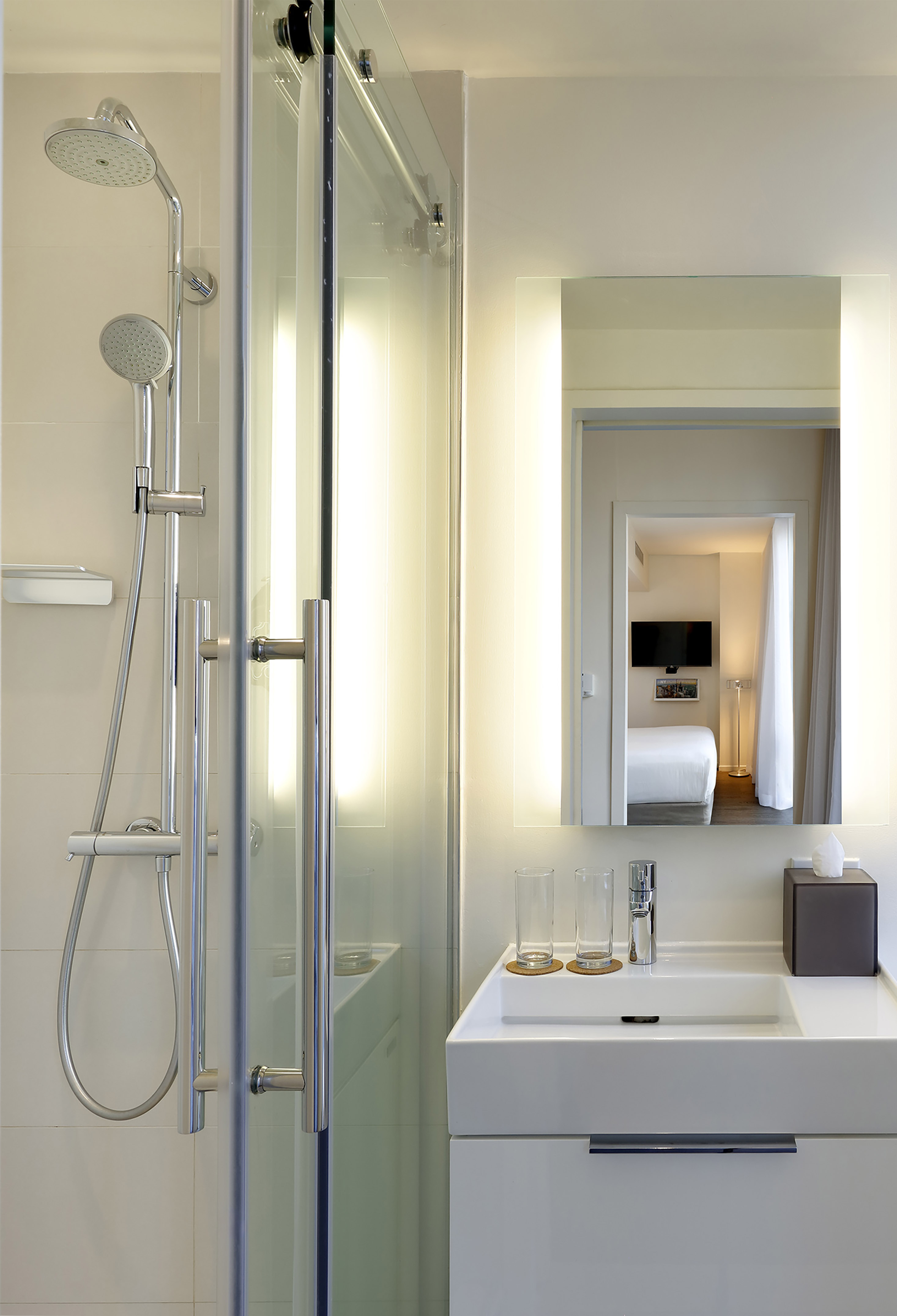 Modern hotel bathroom: glass shower, white vanity, lit mirror reflecting bedroom.