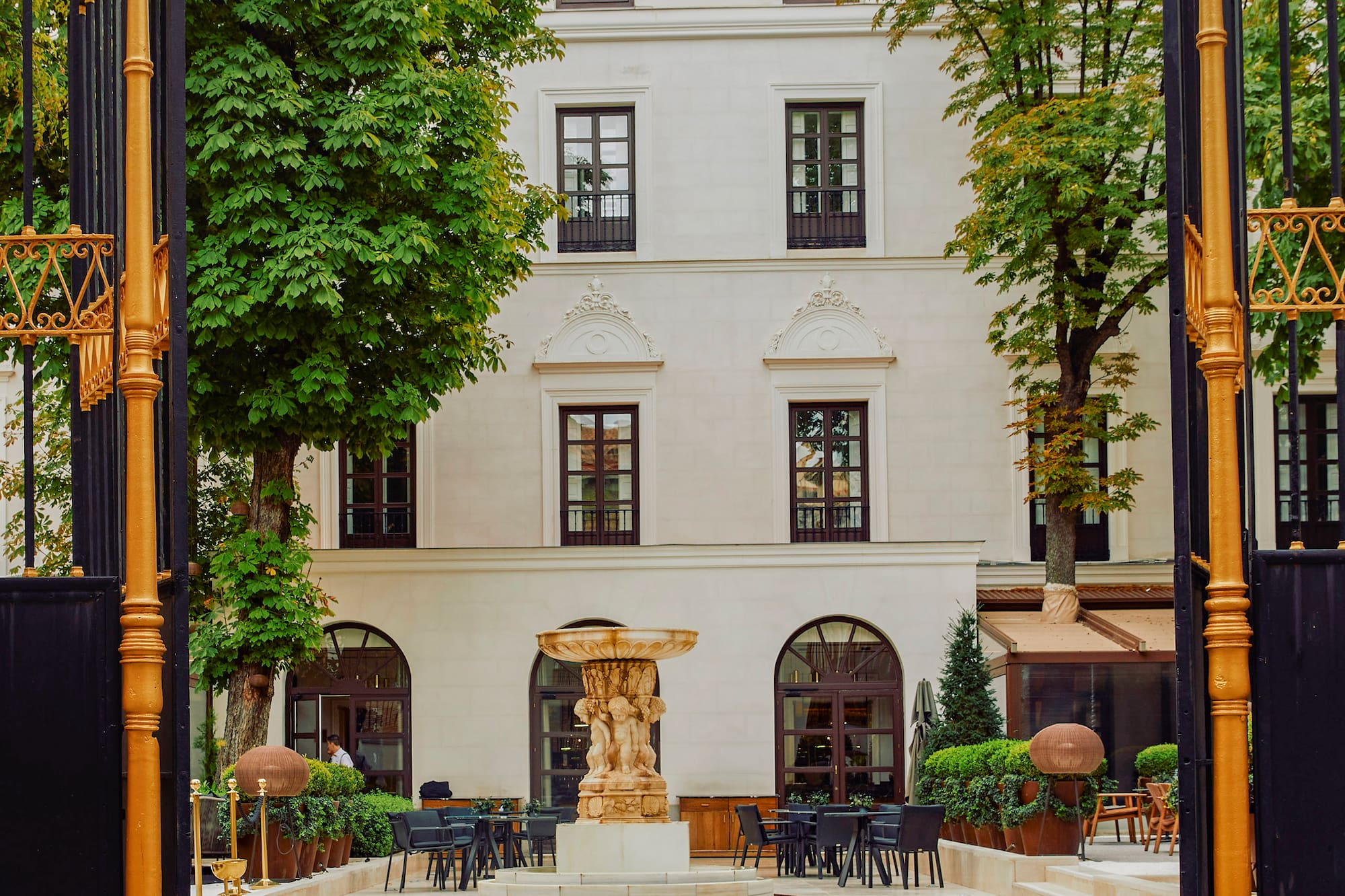 Ornate golden gate opens to building courtyard with fountain, outdoor cafe seating.