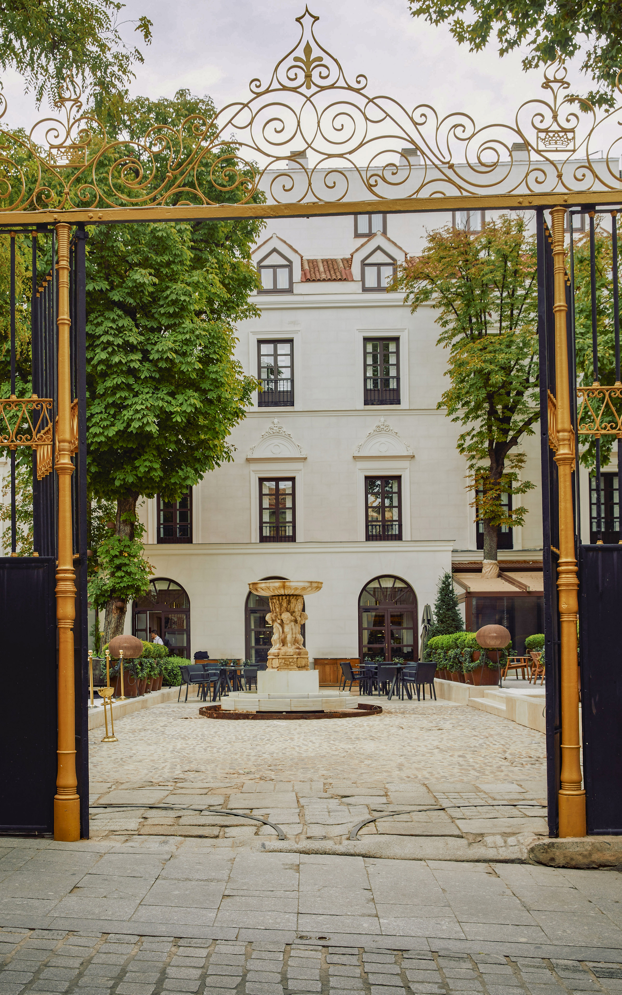 Ornate golden gate opens to building courtyard with fountain, outdoor cafe seating.