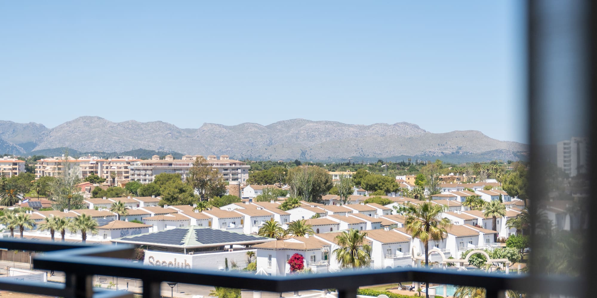 Elevated view of white villas, palm trees, and mountains against a clear sky.