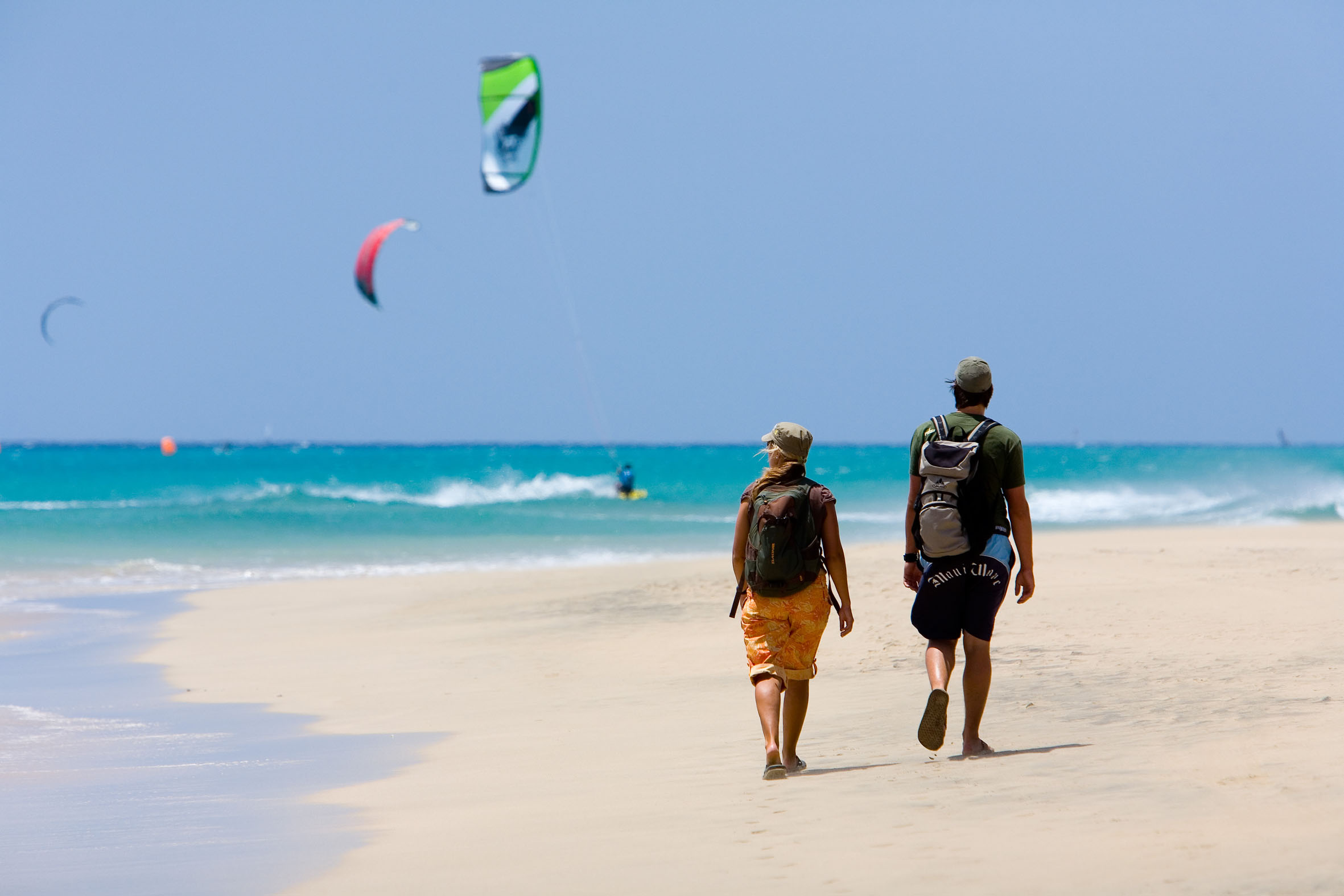 Pareja camina por playa de arena. Mar turquesa con kitesurf y cometas volando.