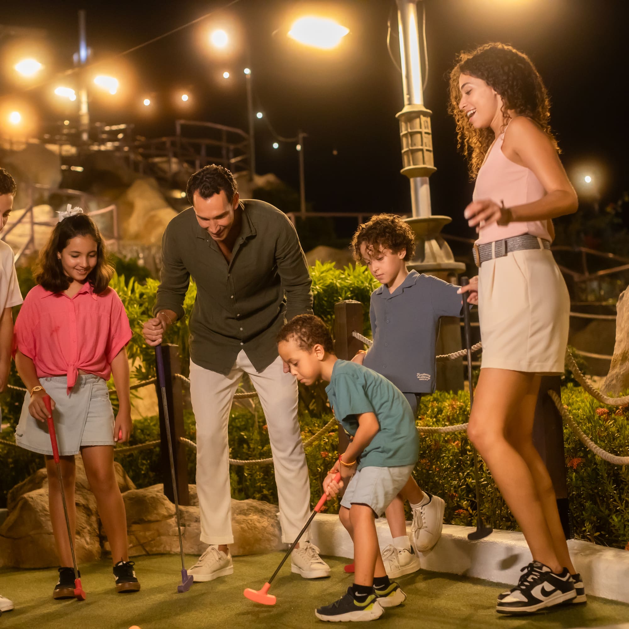 Parents and kids playing night mini golf, smiling on a well-lit course.