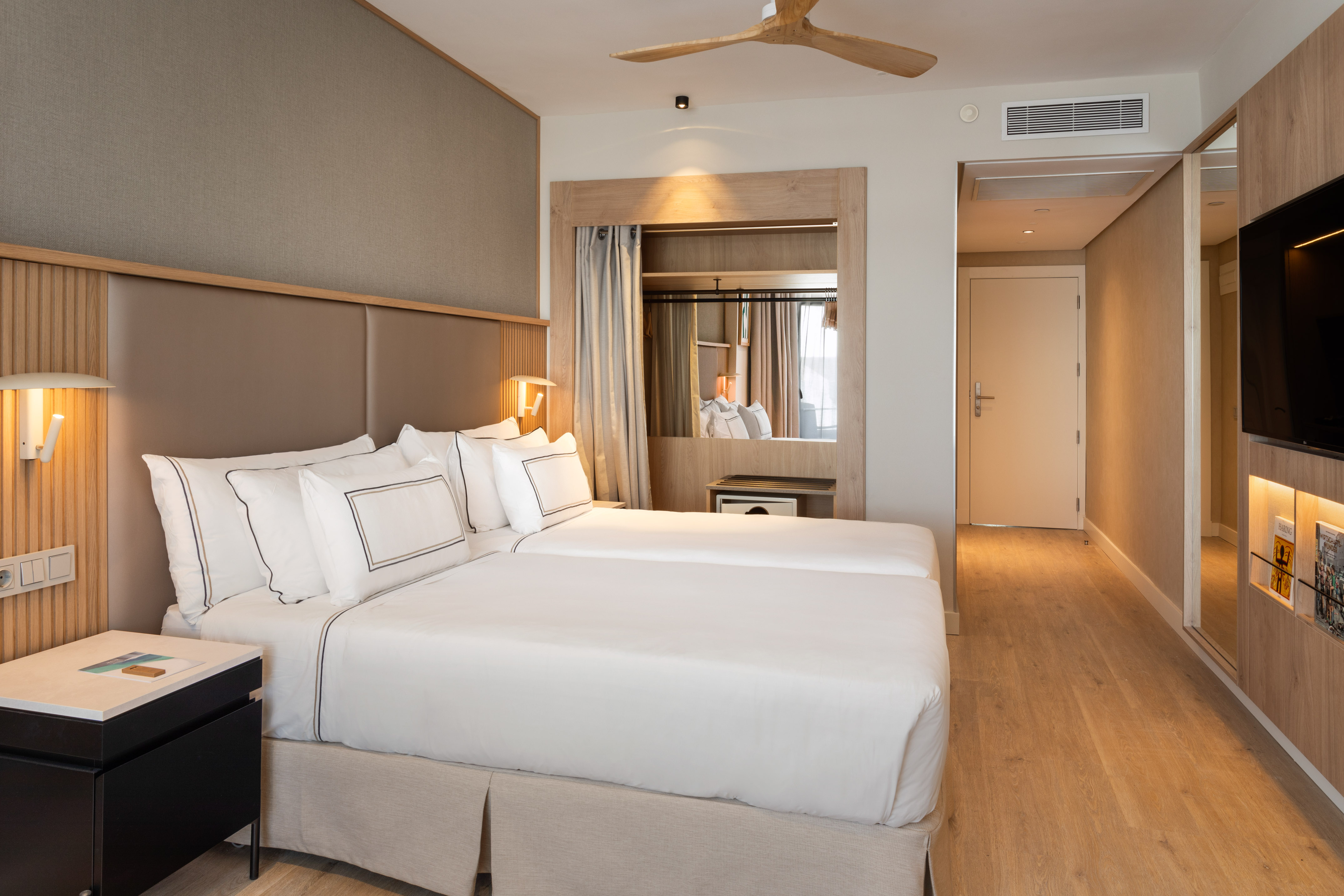 Modern hotel room with twin beds, wooden accents, and ceiling fan.
