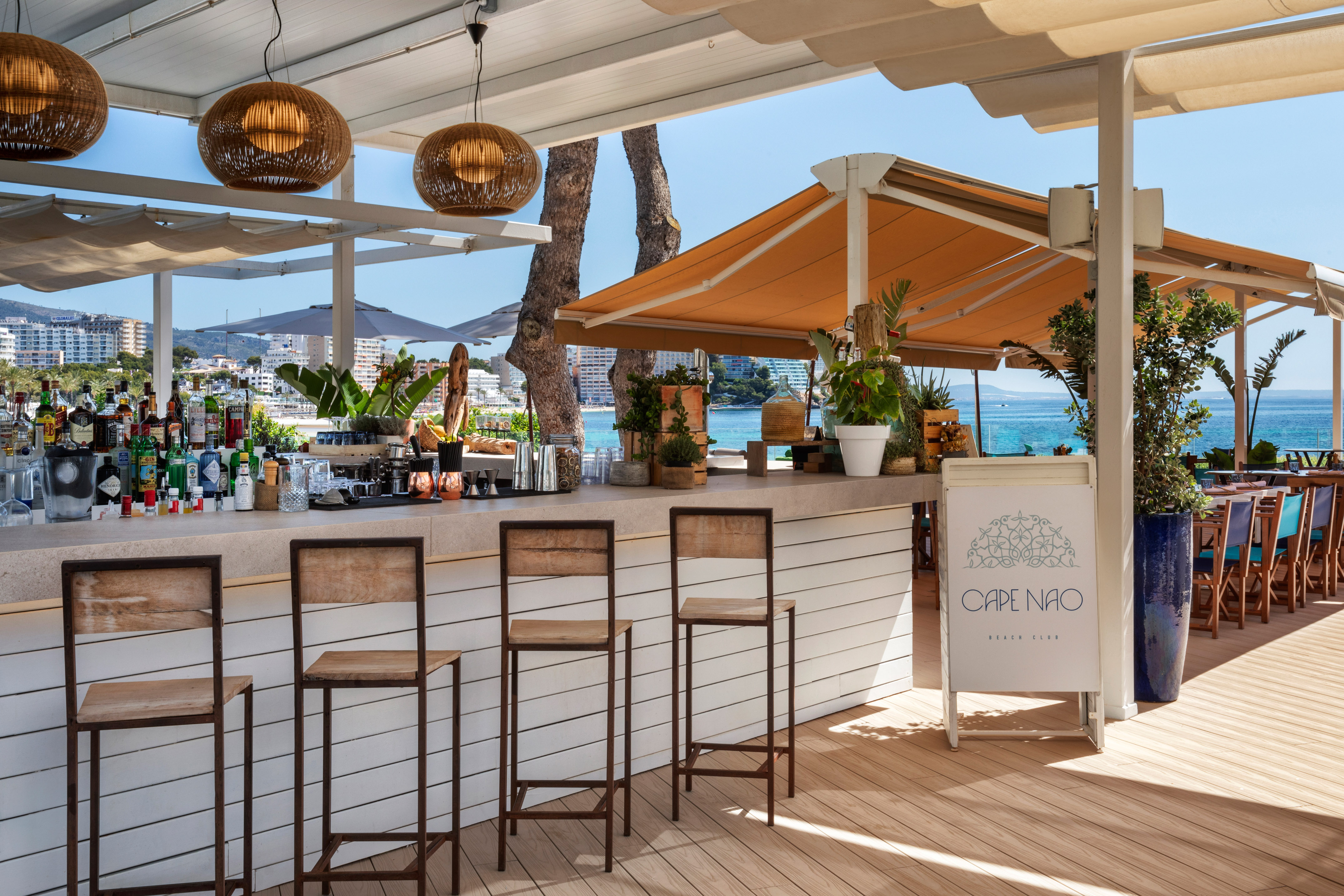 Outdoor bar at Cape Nao Beach Club, ocean view, with wooden stools and bottles.