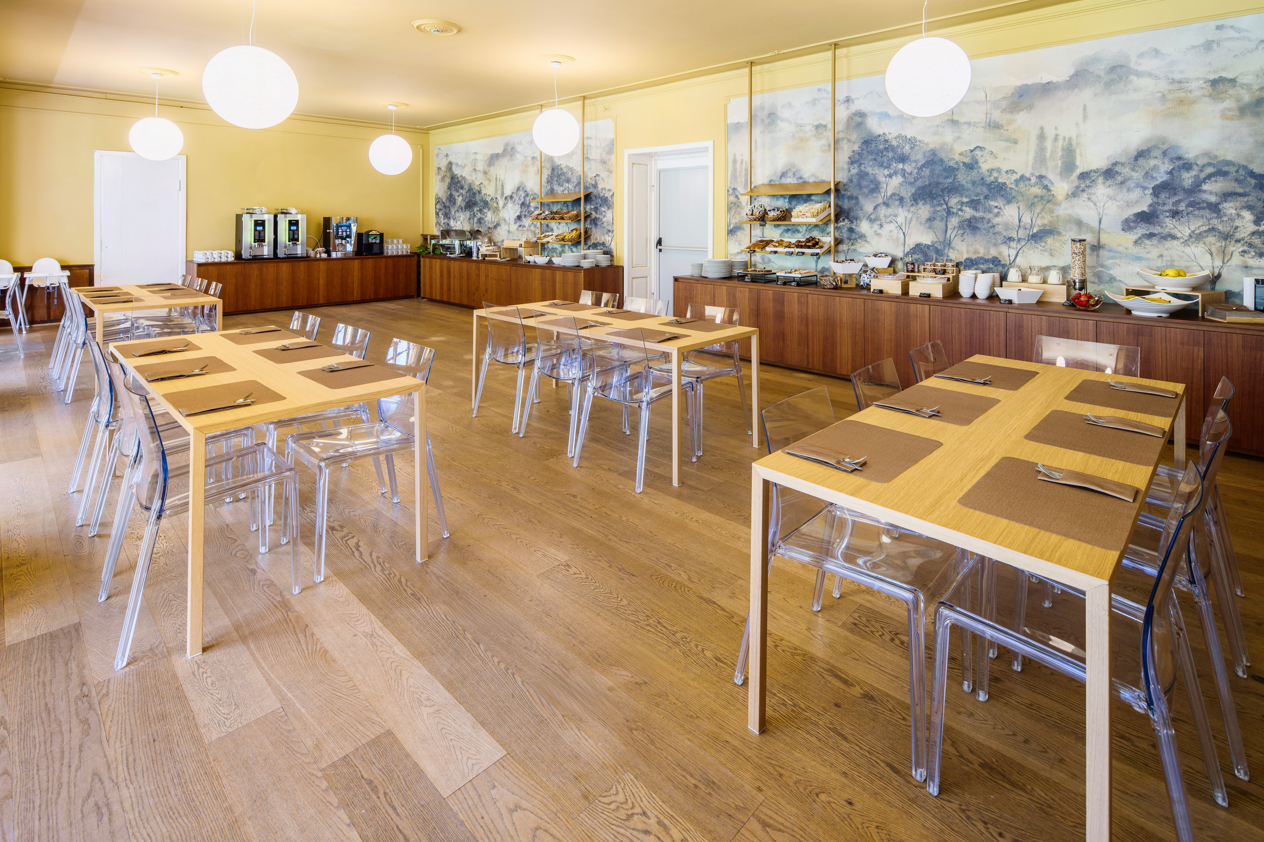 Bright hotel breakfast room, buffet, wooden tables, clear chairs, wall mural.