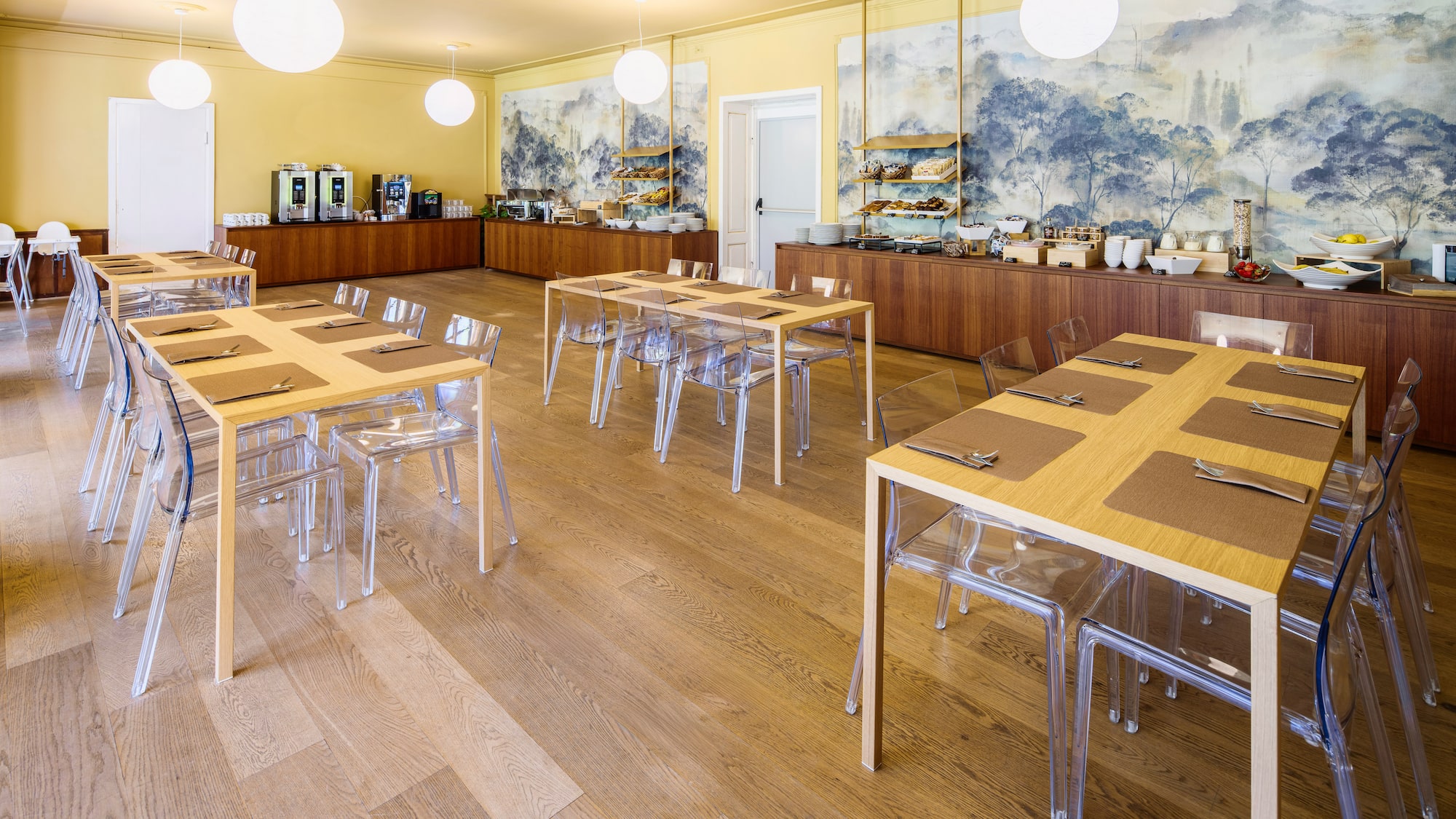 Bright hotel breakfast room, buffet, wooden tables, clear chairs, wall mural.