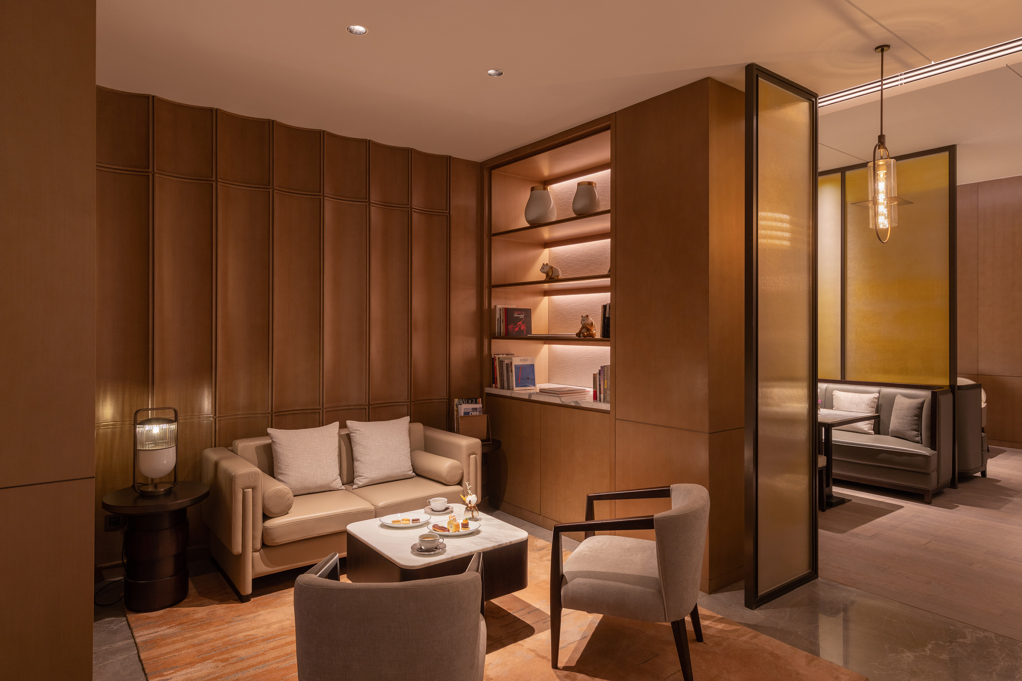 Warm modern lounge with beige seating, coffee table, and illuminated bookshelf.