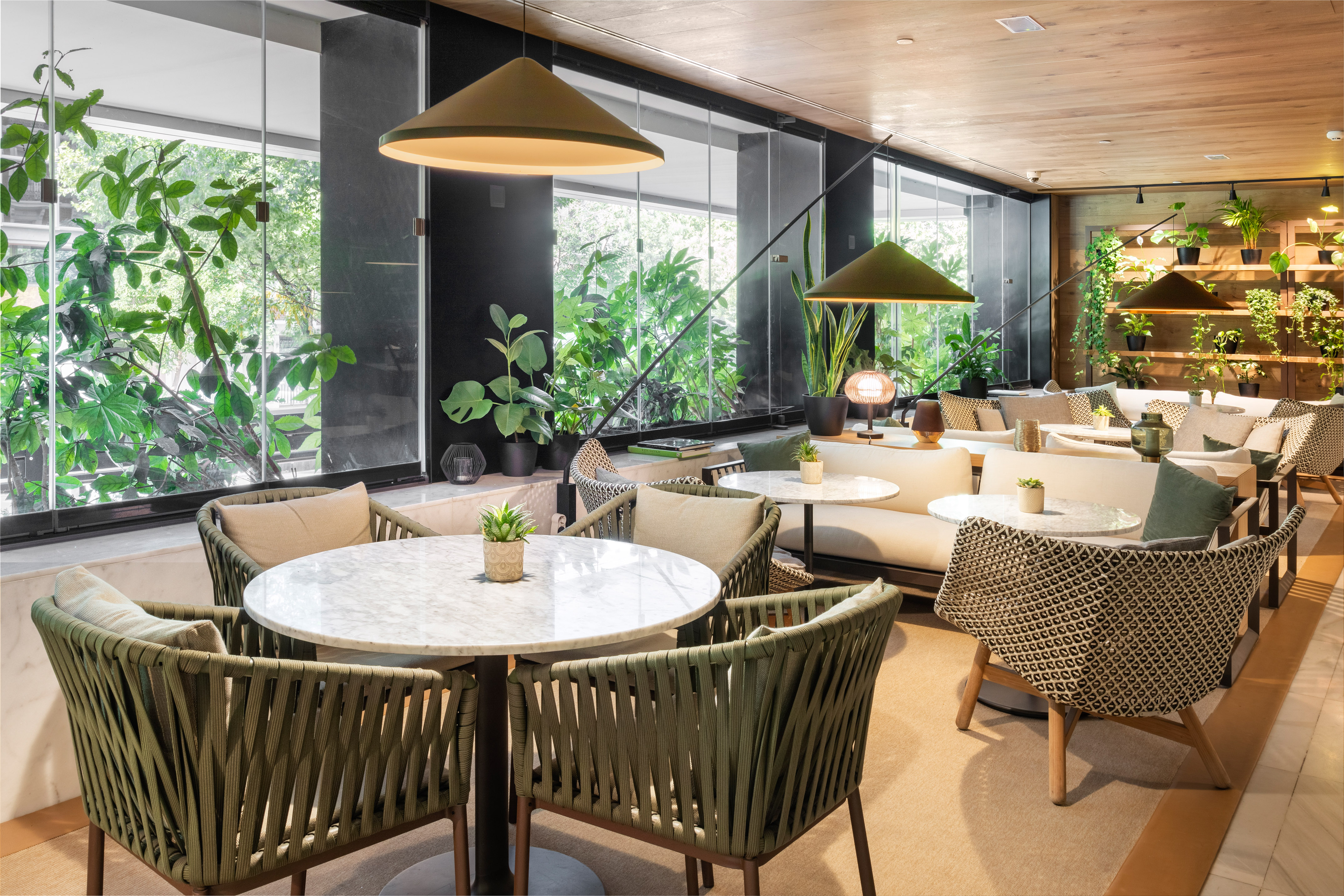 Biophilic lounge interior with green plants, comfy seating, marble tables, and large windows.