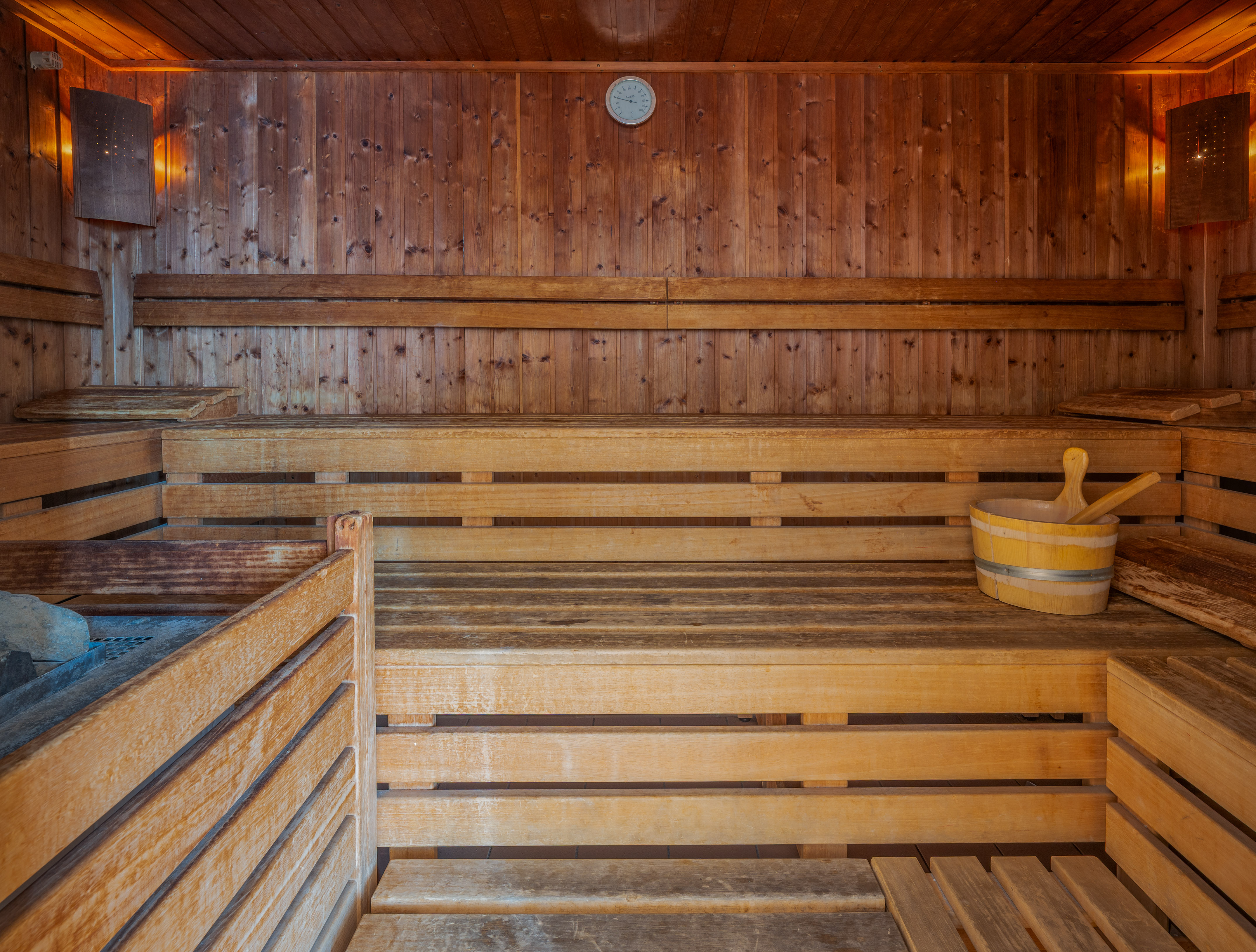 Wooden sauna interior with multiple benches, heater, bucket, and thermometer.