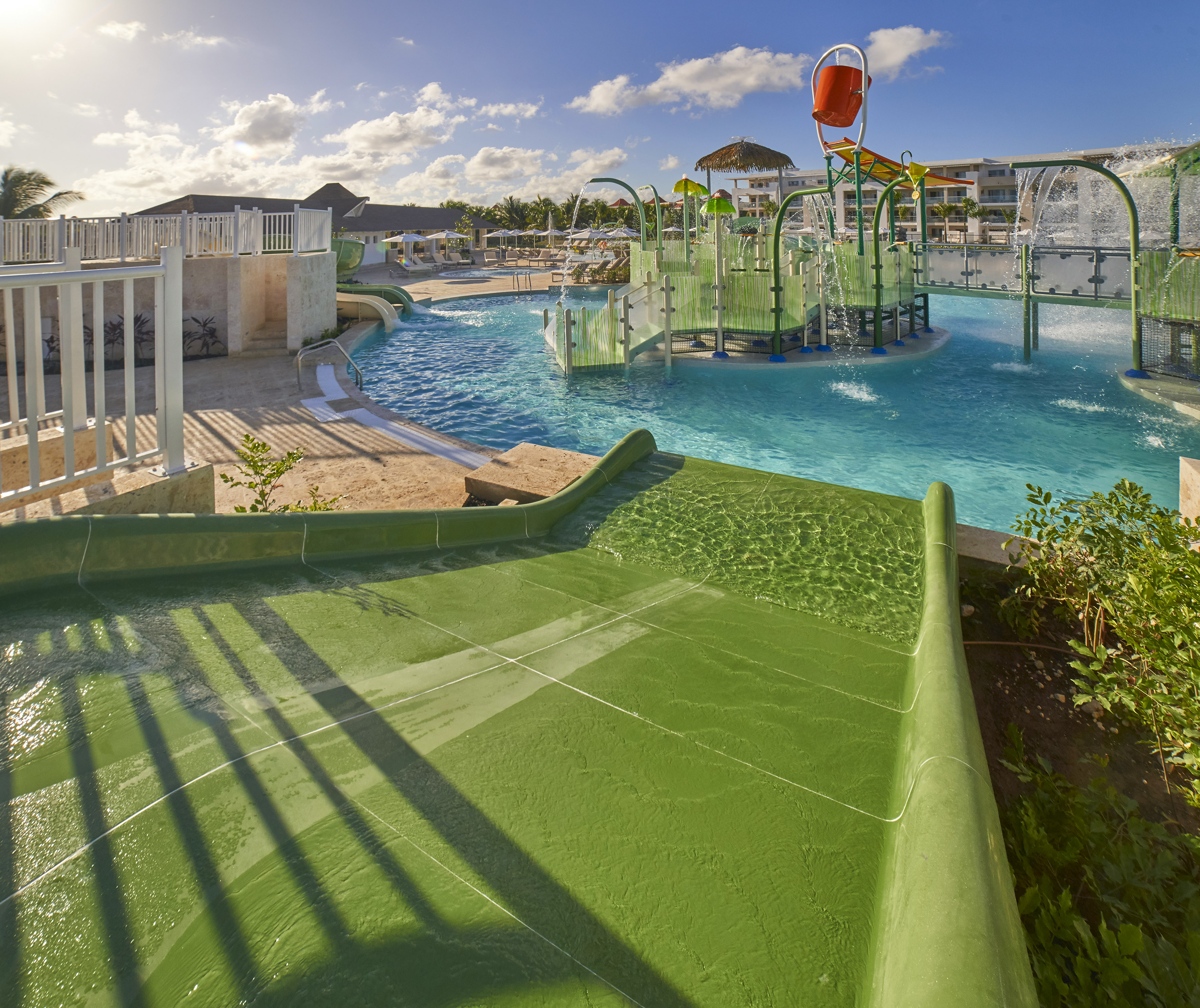 Sunny resort water park with green slides, blue pools, and splashing features.
