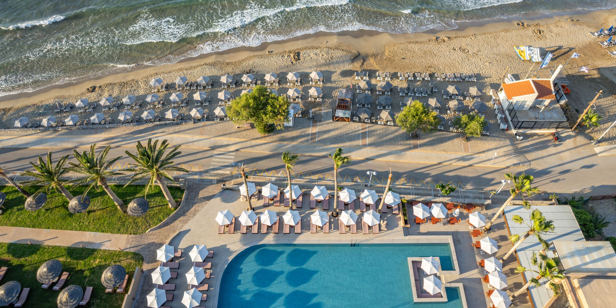 Aerial view: Beach hotel with ocean, sandy shore, pool, umbrellas, palm trees.