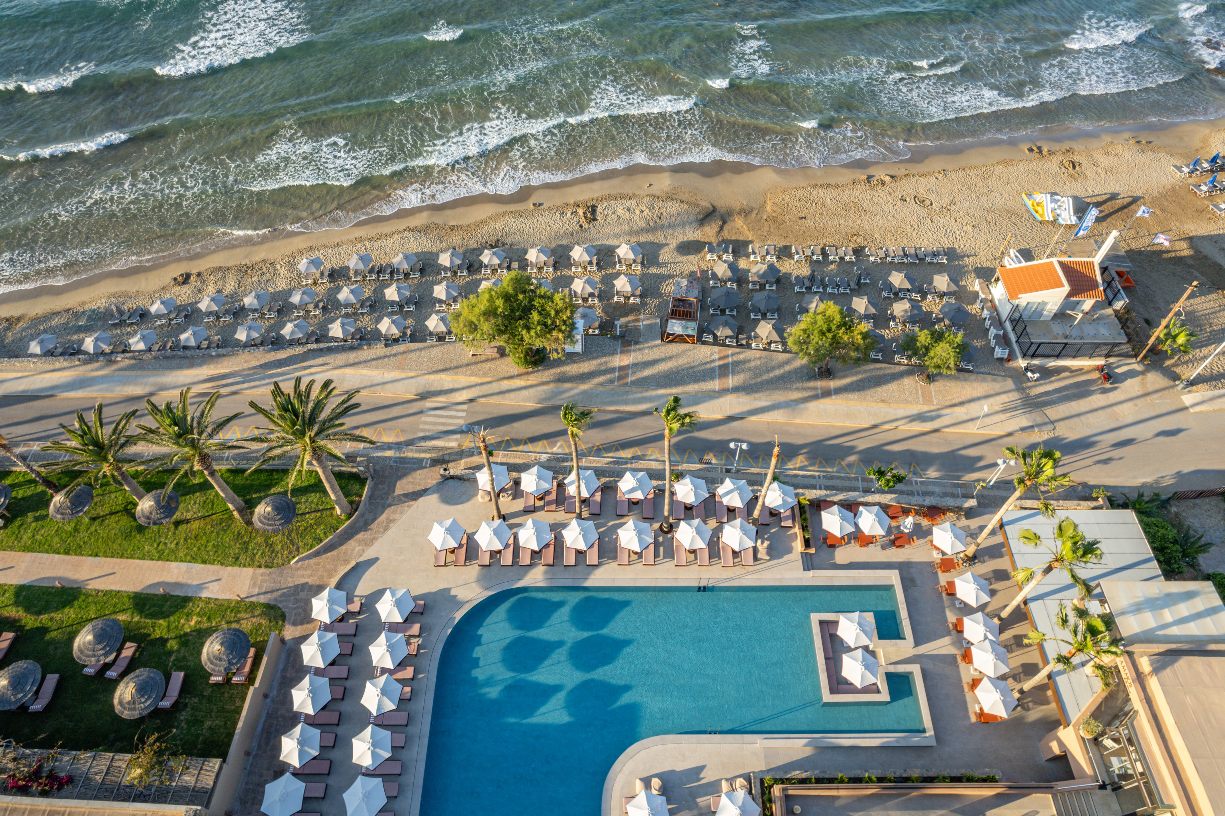 Aerial view: Beach hotel with ocean, sandy shore, pool, umbrellas, palm trees.