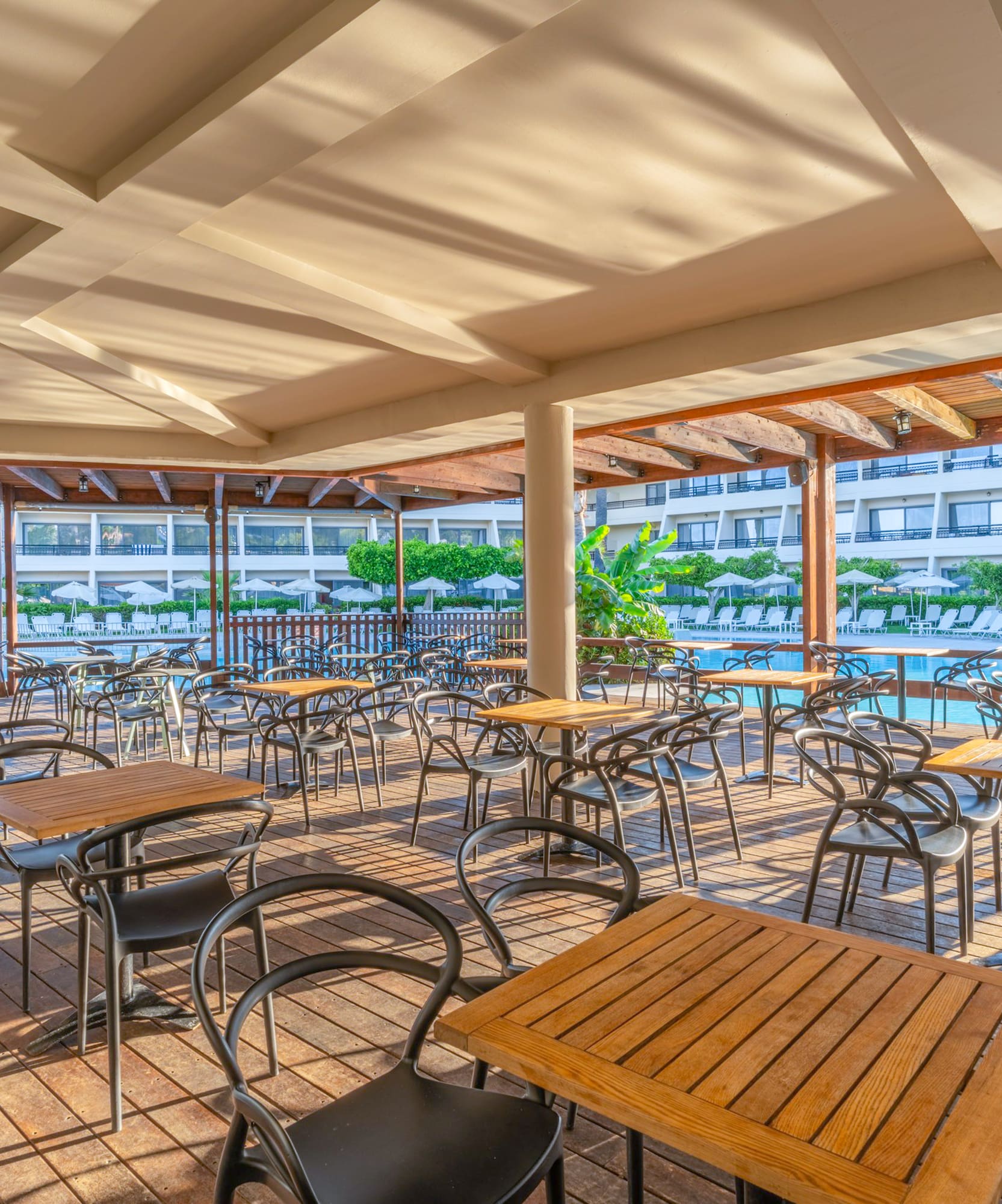 Sunny outdoor dining terrace overlooking a hotel pool with tables and chairs.