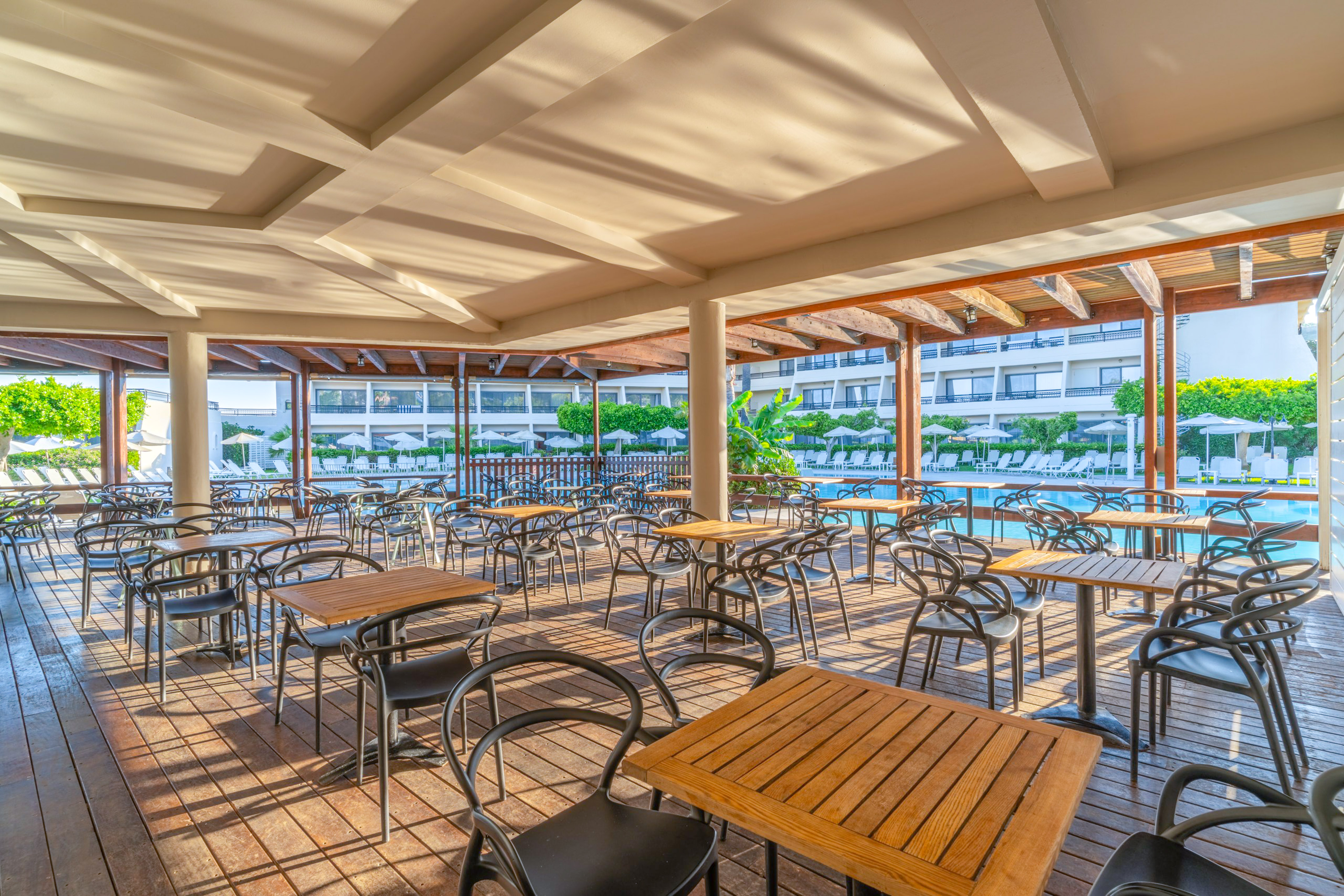 Sunny outdoor dining terrace overlooking a hotel pool with tables and chairs.