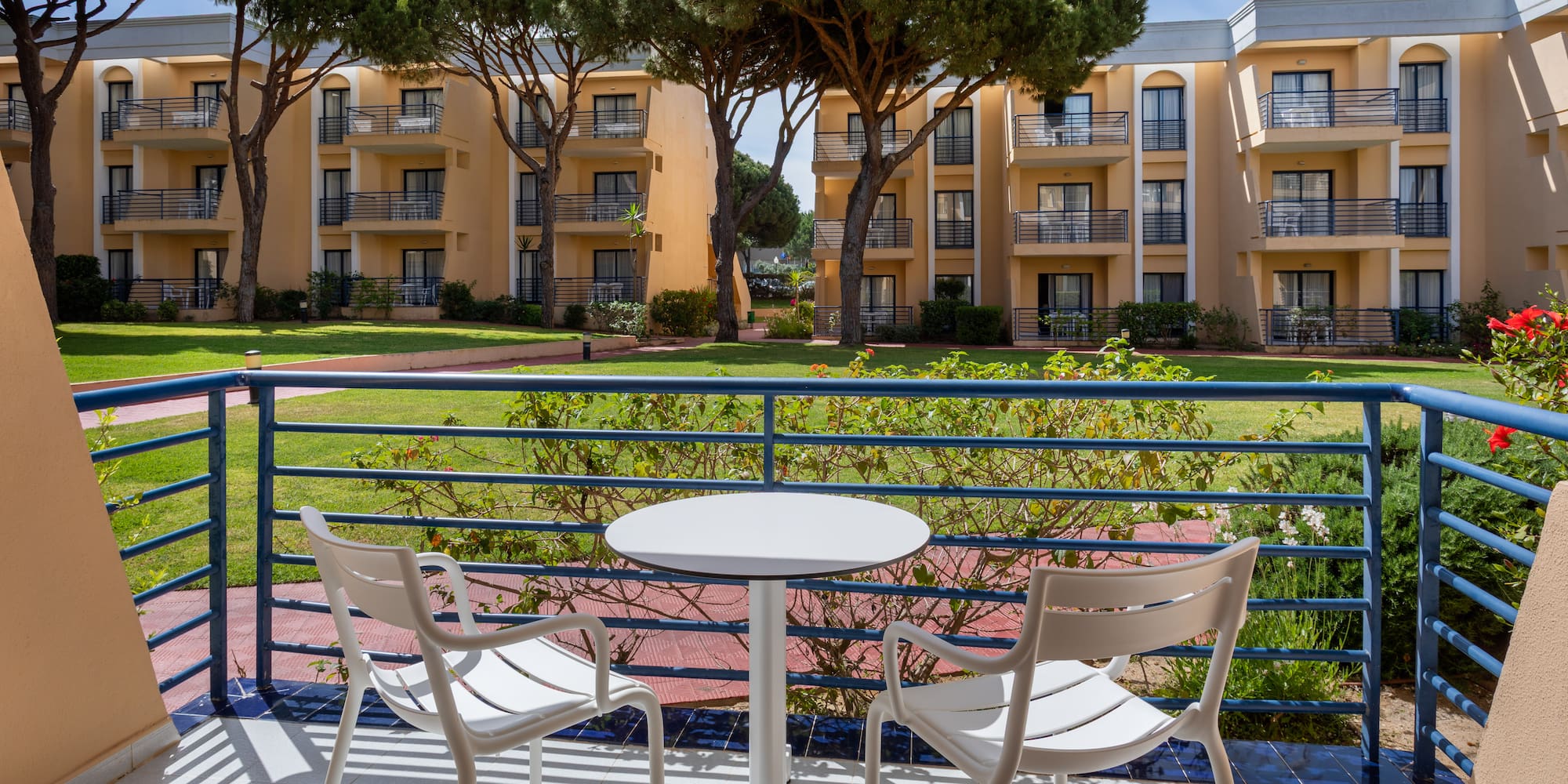 Balcony with outdoor seating, garden, and hotel buildings with pine trees.