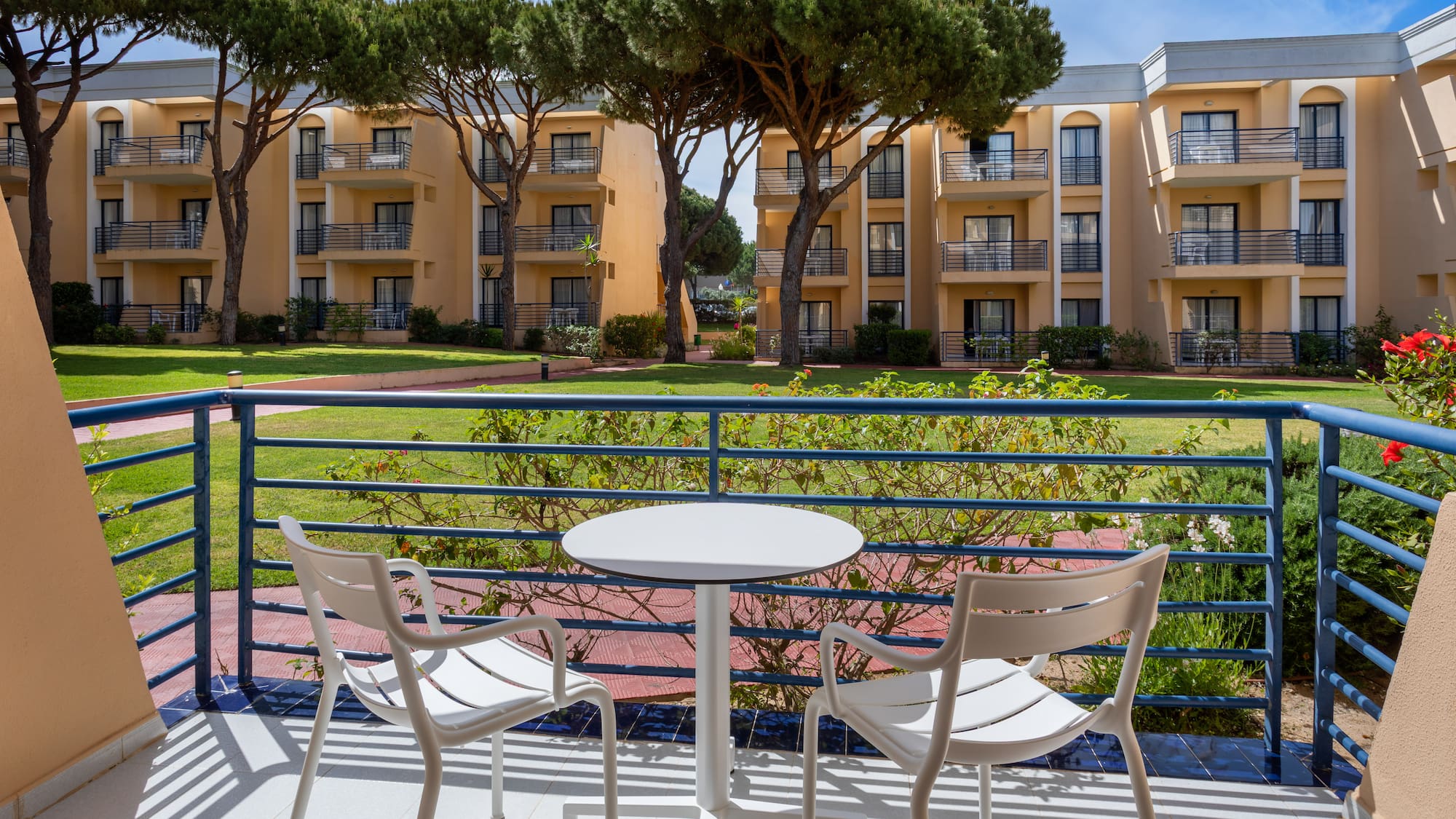 Sunny hotel balcony with white table, chairs, resort garden, and apartment buildings.