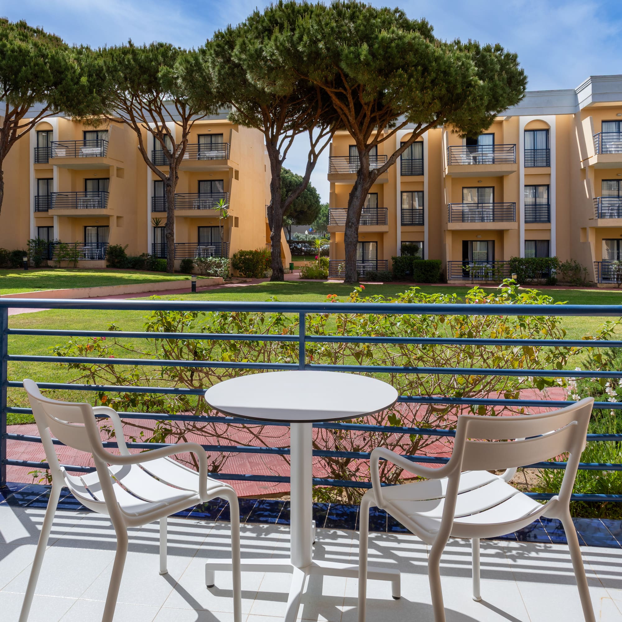 Balcony with outdoor seating, garden, and hotel buildings with pine trees.