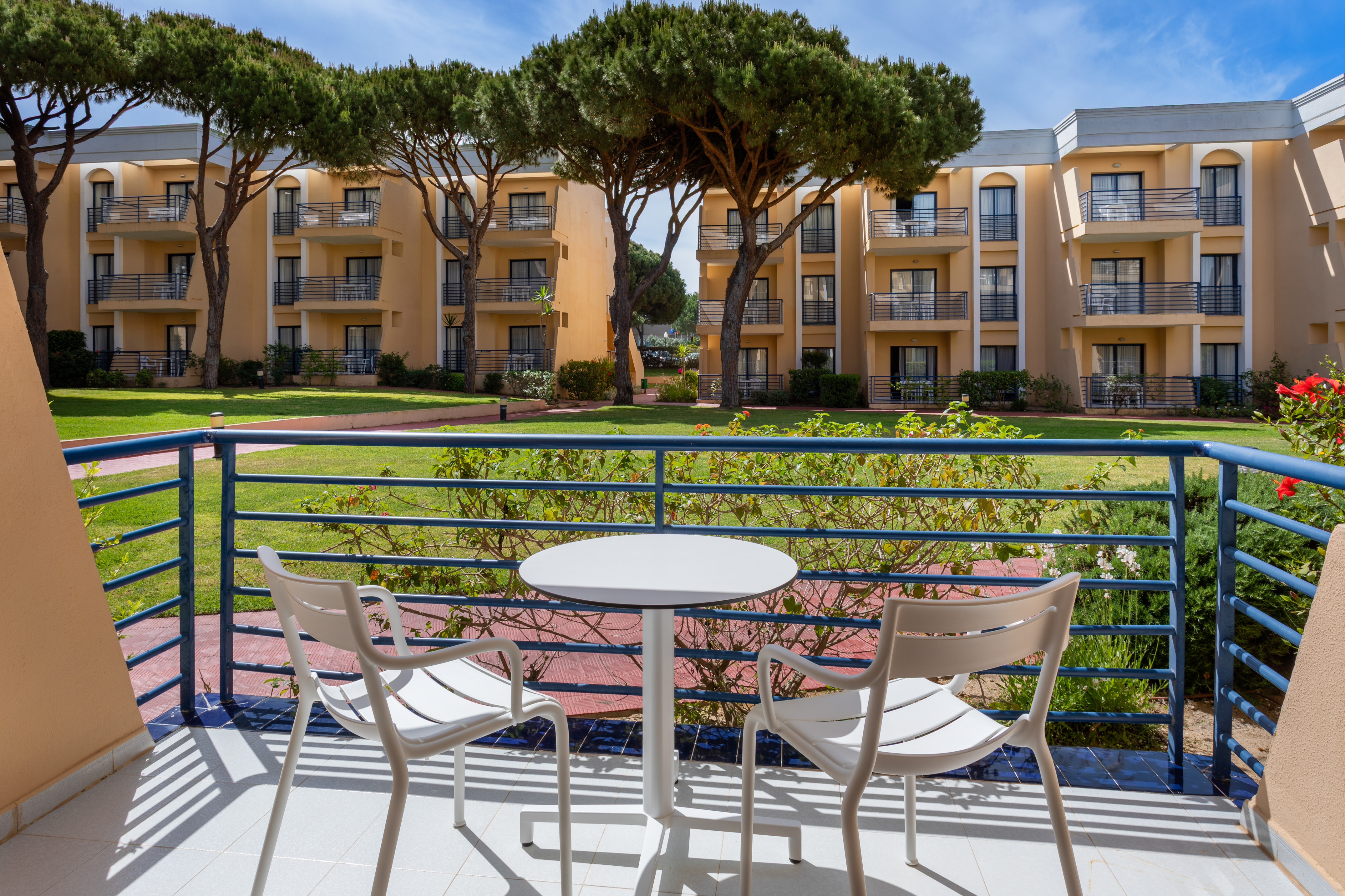 Sunny hotel balcony with white table, chairs, resort garden, and apartment buildings.
