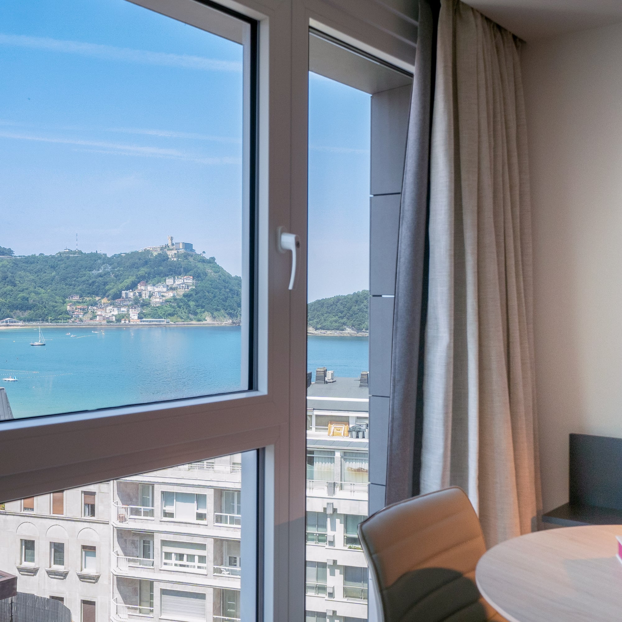 Vista desde ventana a la Bahía de La Concha, San Sebastián, con playa y montañas.